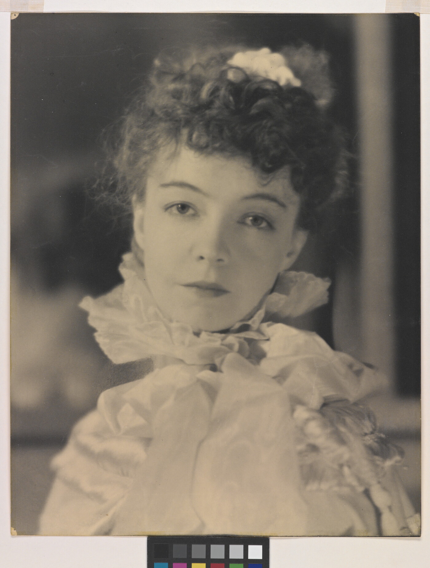 A black-and-white portrait photograph of a White woman with curly hair pulled on top of her head, wearing a top with a ruffled collar.