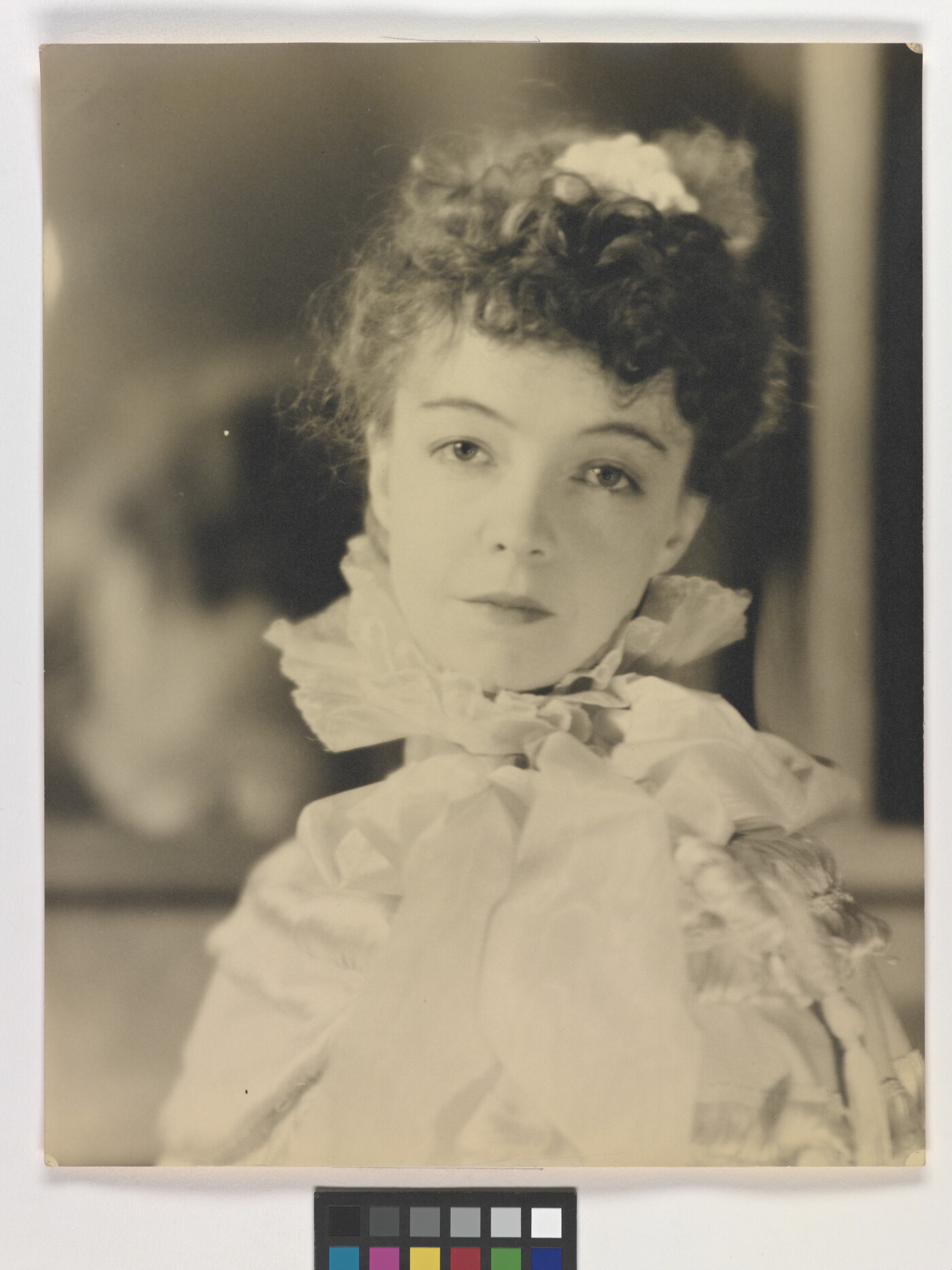 A black-and-white portrait photograph of a White woman with curly hair pulled on top of her head, wearing a top with a ruffled collar.