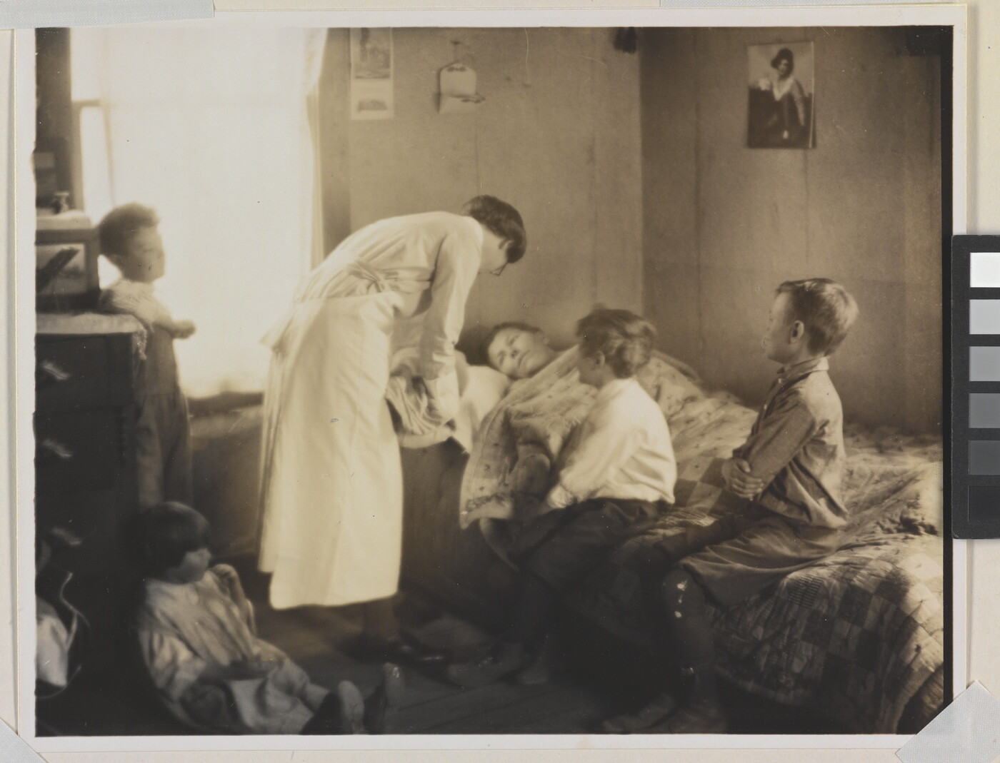 A black-and-white photograph of a nurse bending over a person in a bed, two children sitting on the bed, and two children behind the nurse.