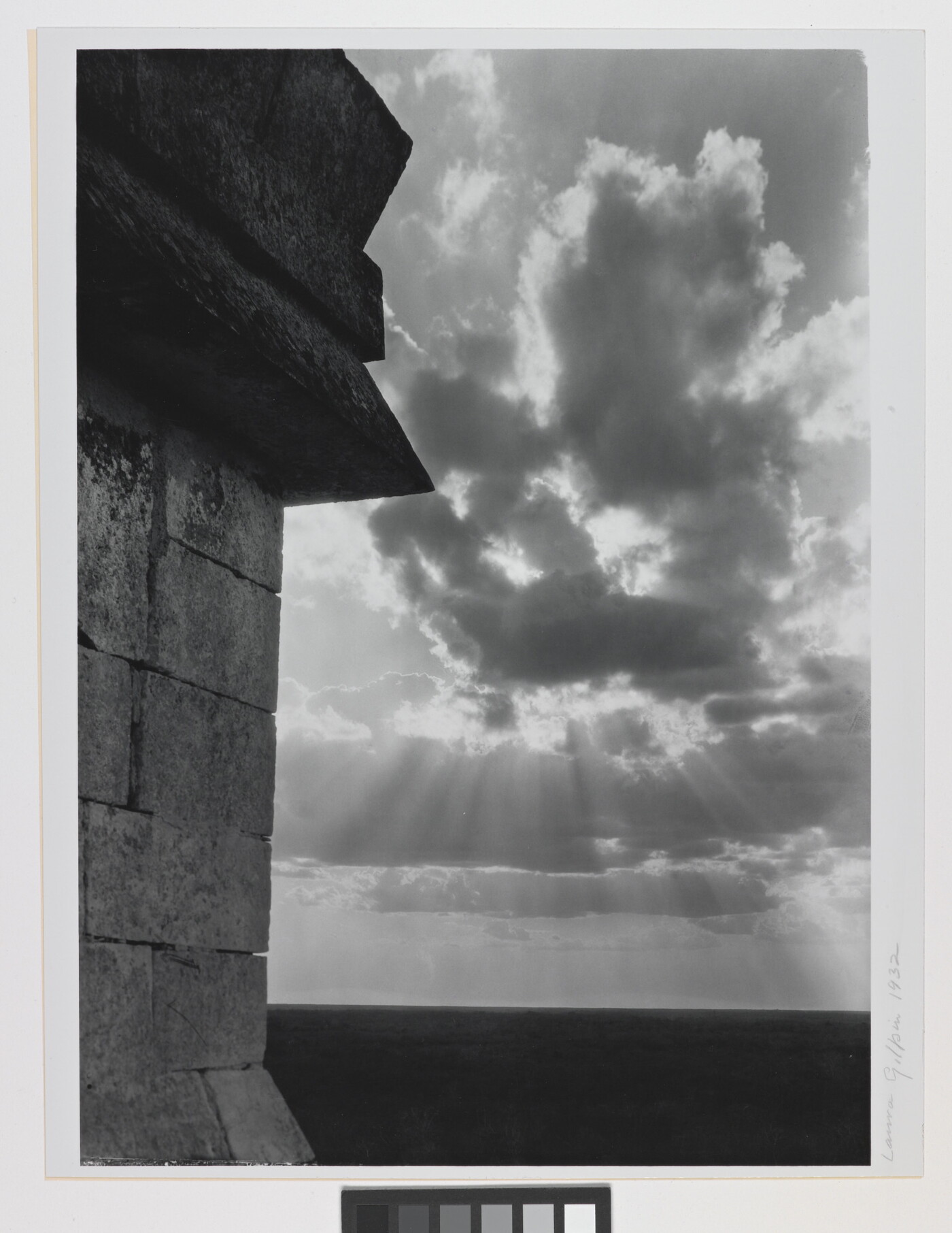 A black-and-white photograph of the sun&#039;s rays bursting from clouds and a stark brick architectural feature in the left foreground.