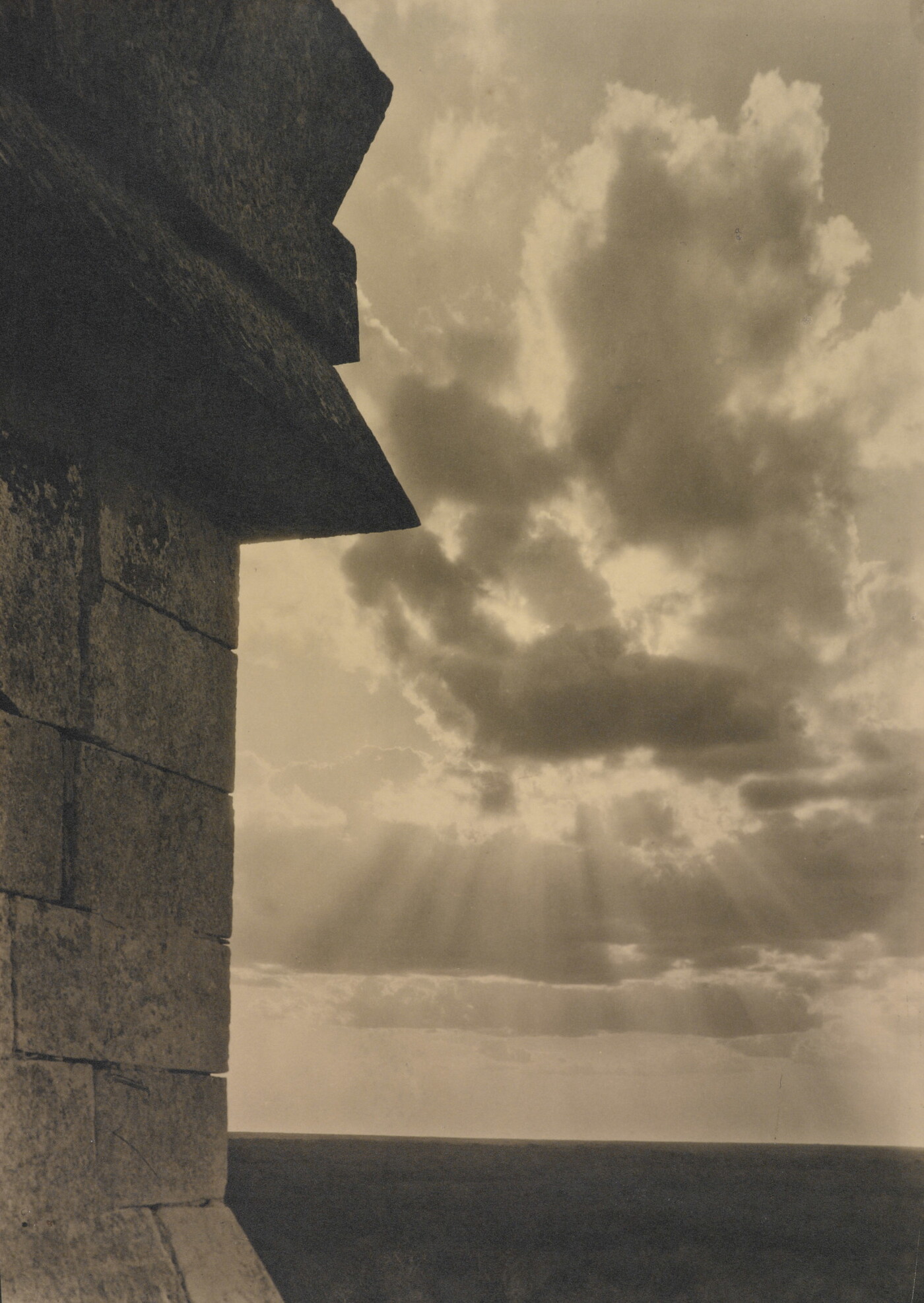 A sepia-toned photograph of the sun&#039;s rays bursting from clouds and a stark brick architectural feature in the left foreground.