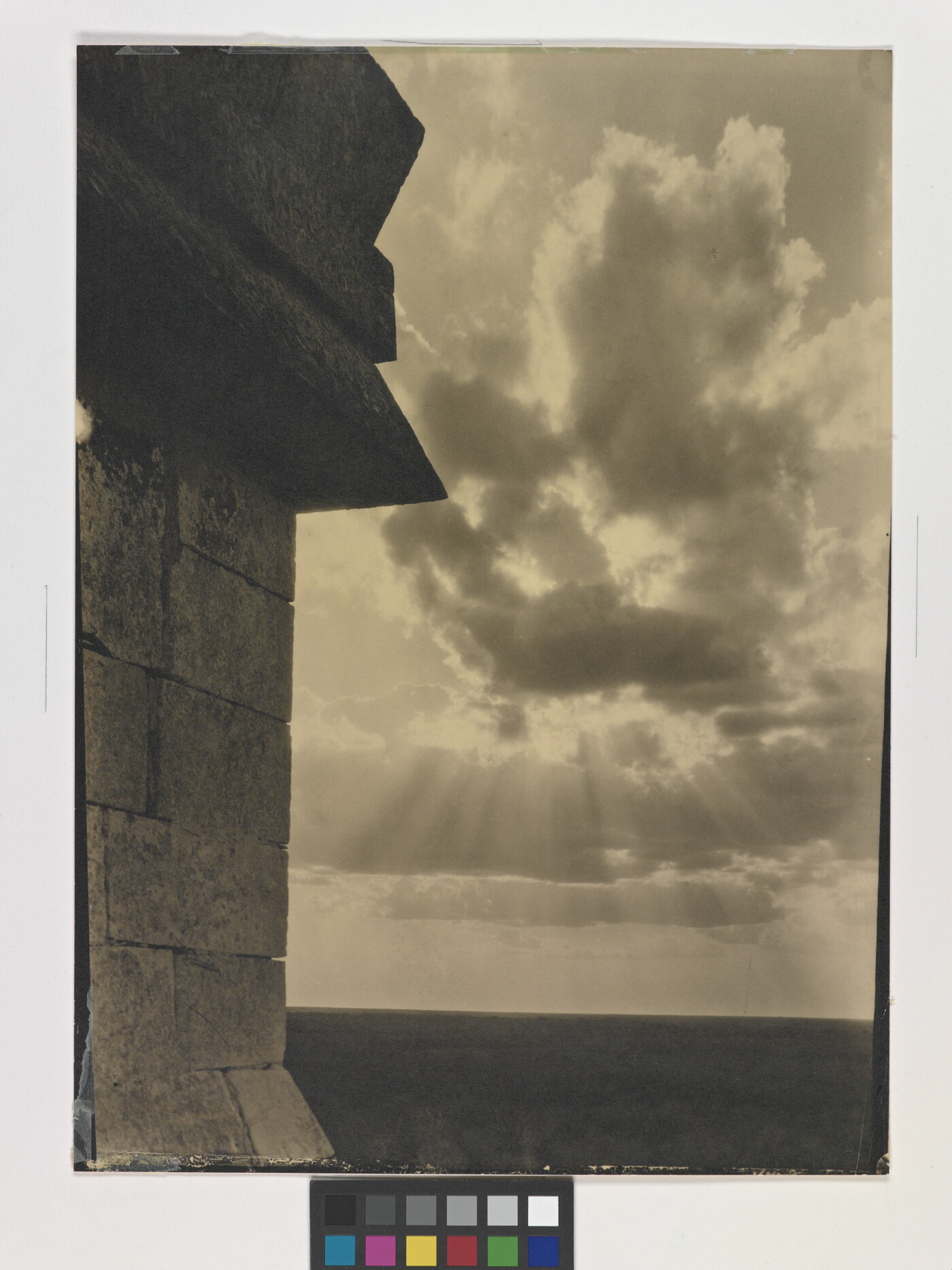 A sepia-toned photograph of the sun&#039;s rays bursting from clouds and a stark brick architectural feature in the left foreground.