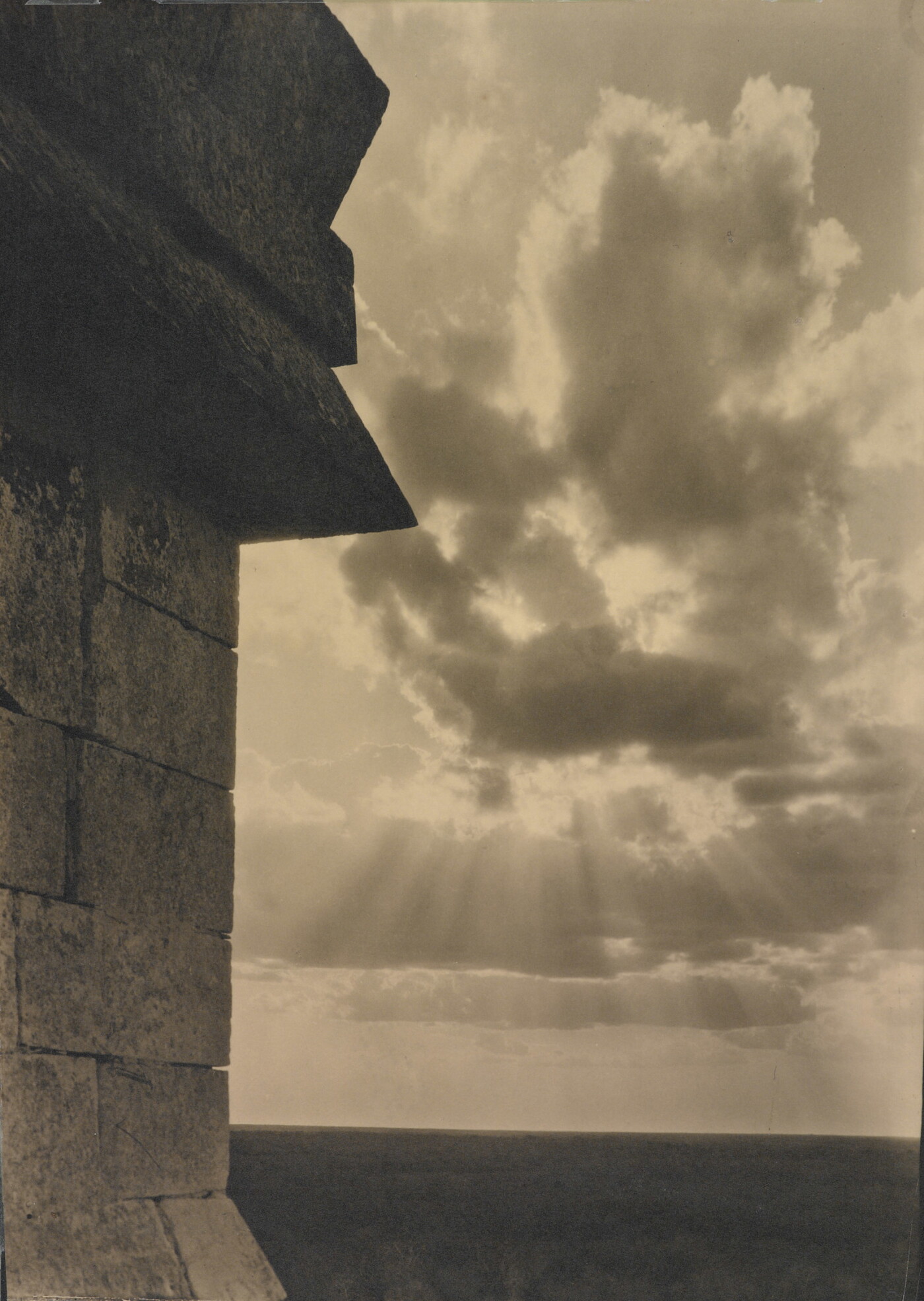 A sepia-toned photograph of the sun's rays bursting from clouds and a stark brick architectural feature in the left foreground.