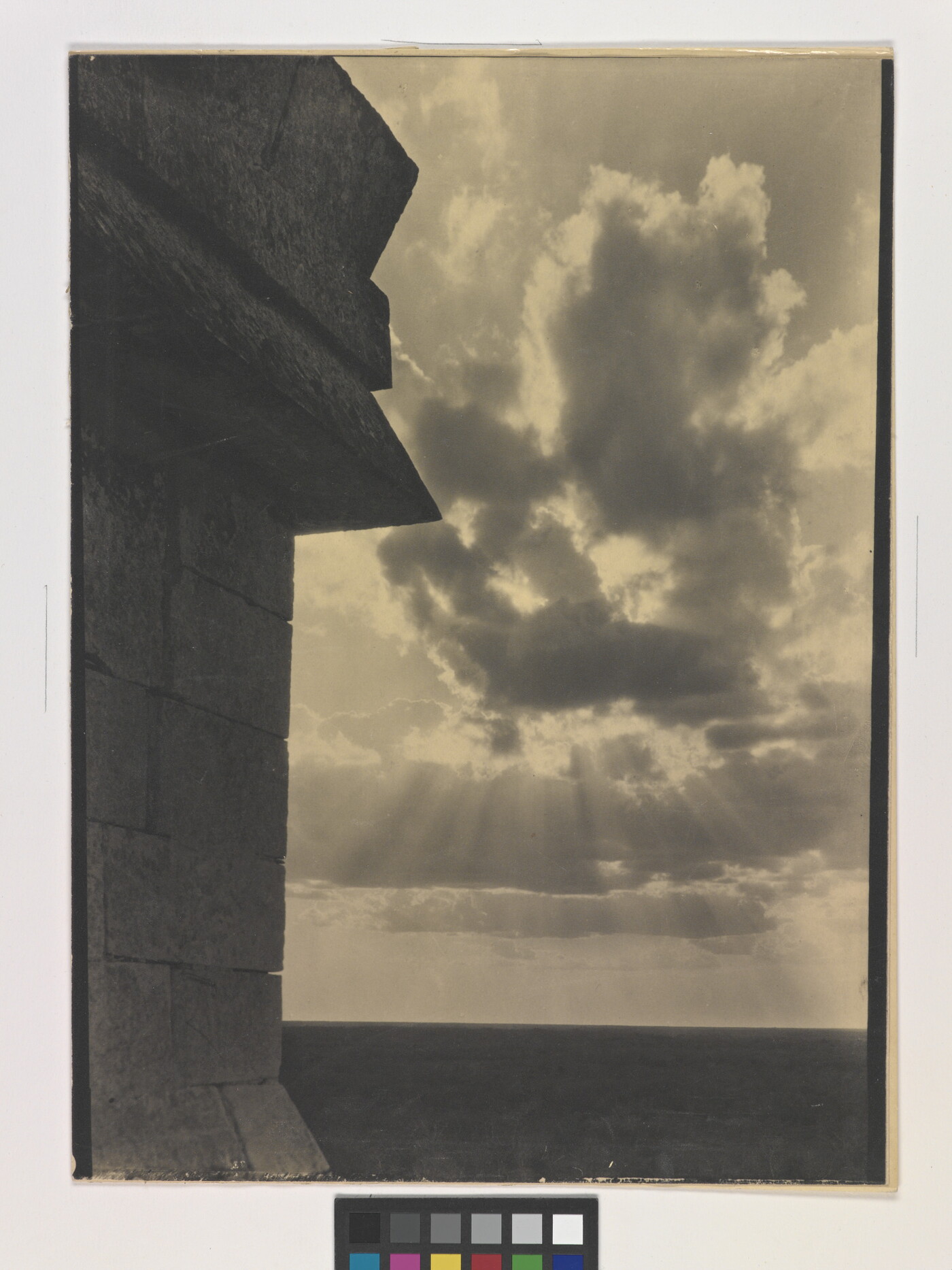 A sepia-toned photograph of the sun&#039;s rays bursting from clouds and a stark brick architectural feature in the left foreground.