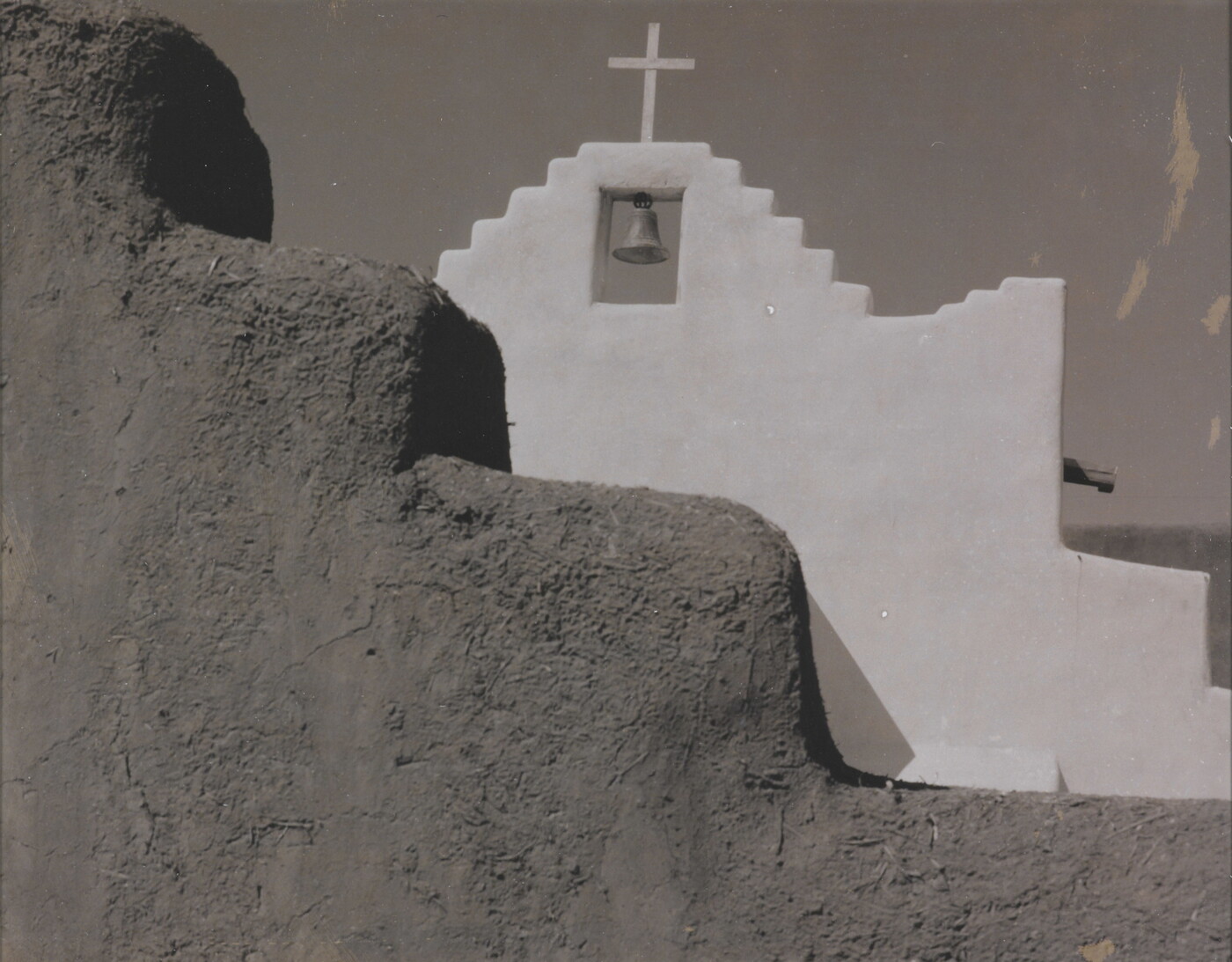 A black-and-white photograph of a terraced adobe bell tower with a cross on top, partially hidden by a terraced adobe wall.
