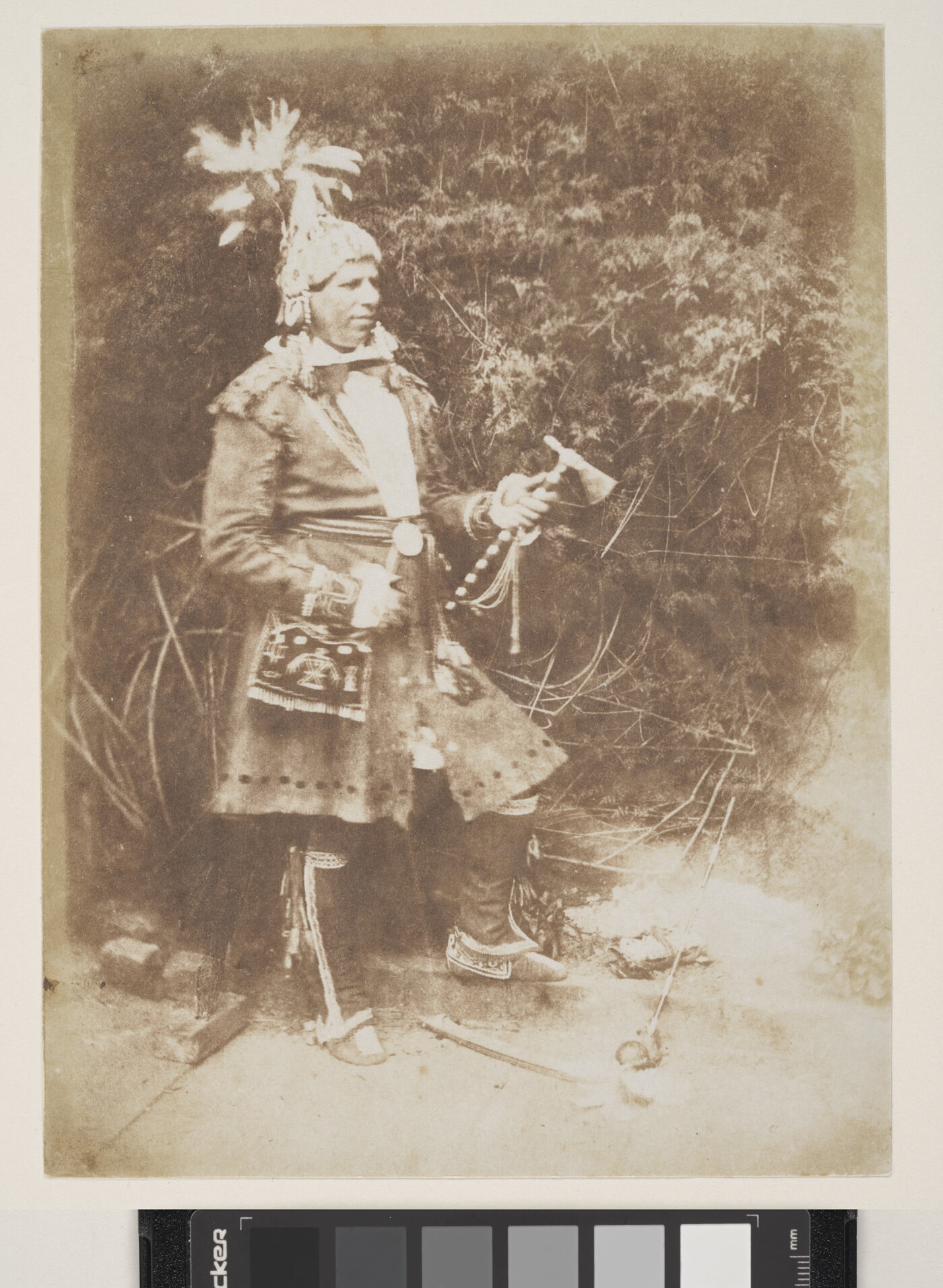 A sepia-toned photograph of an Indigenous man wearing traditional clothing and holding a tomahawk.