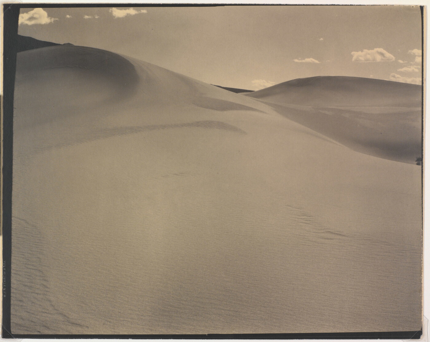 A sepia-toned photograph of a rippling sand dune rising up into a sky dotted with a few small clouds.