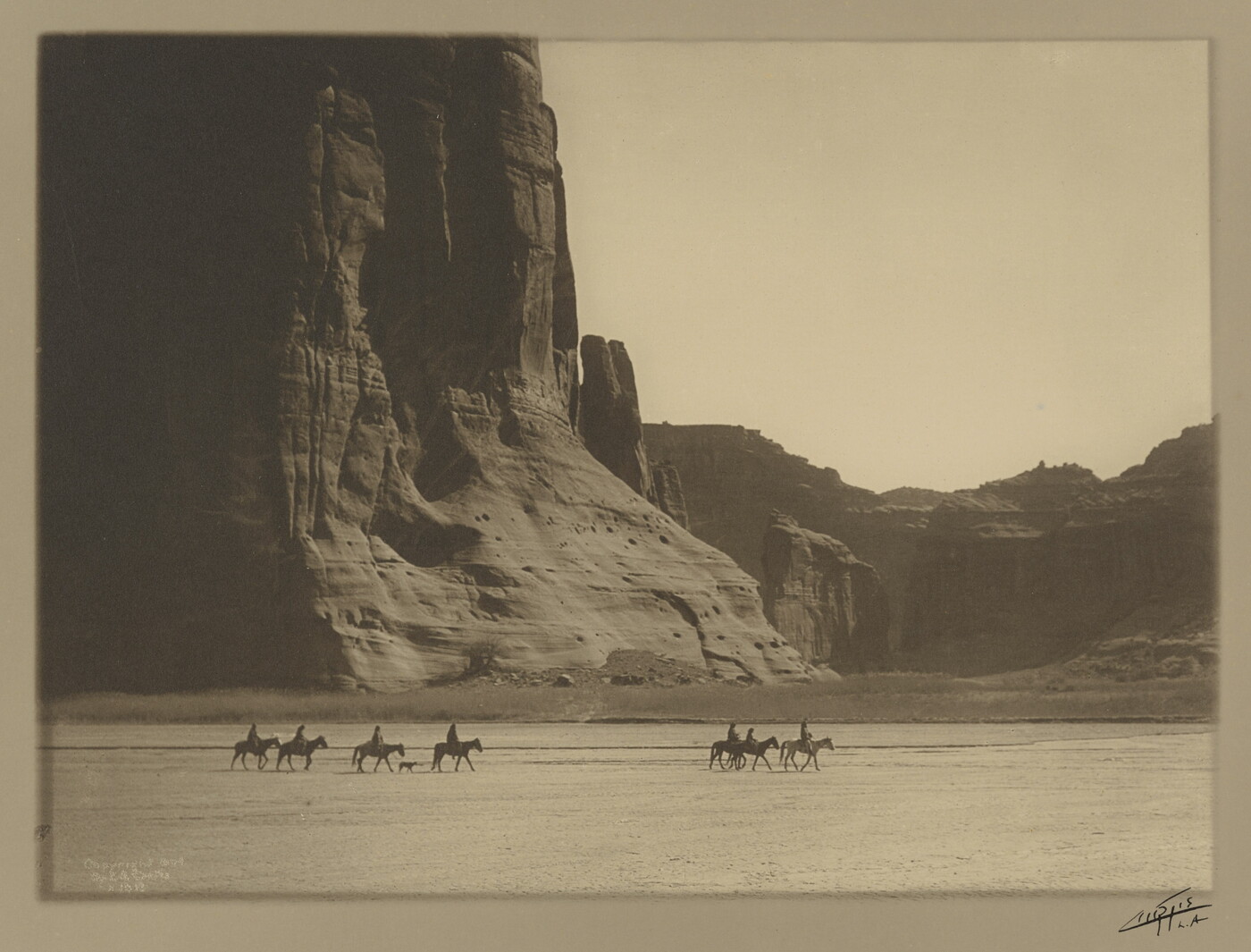 A sepia-toned photograph of a group of people on horses dwarfed by the towering canyon and rock formations behind them.