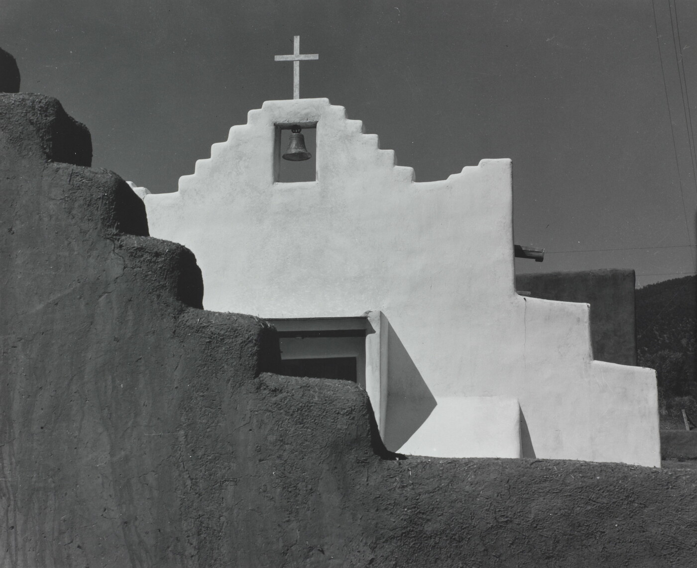 A black-and-white photograph of a terraced adobe bell tower with a cross on top, partially hidden by a terraced adobe wall.