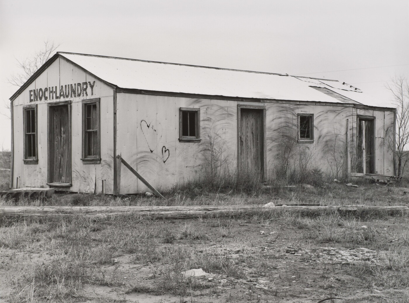 A black-and-white photograph of a one-story building with boarded up windows and graffiti on the walls; lettering above the door reads, "Enoch Laundry."