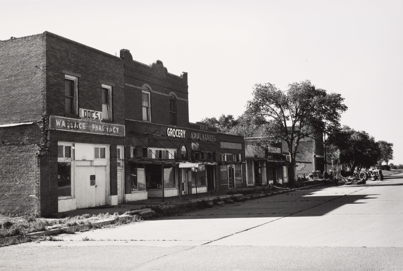 A black-and-white photograph of a paved street lined with brick buildings, some boarded up, casting stark shadows on the ground.