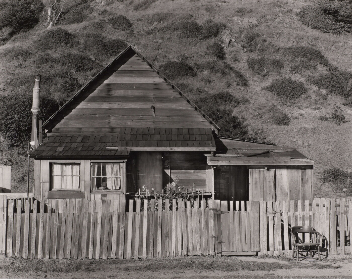 A black-and-white photograph of a small clapboard house behind a picket fence with a chair next to the gate.
