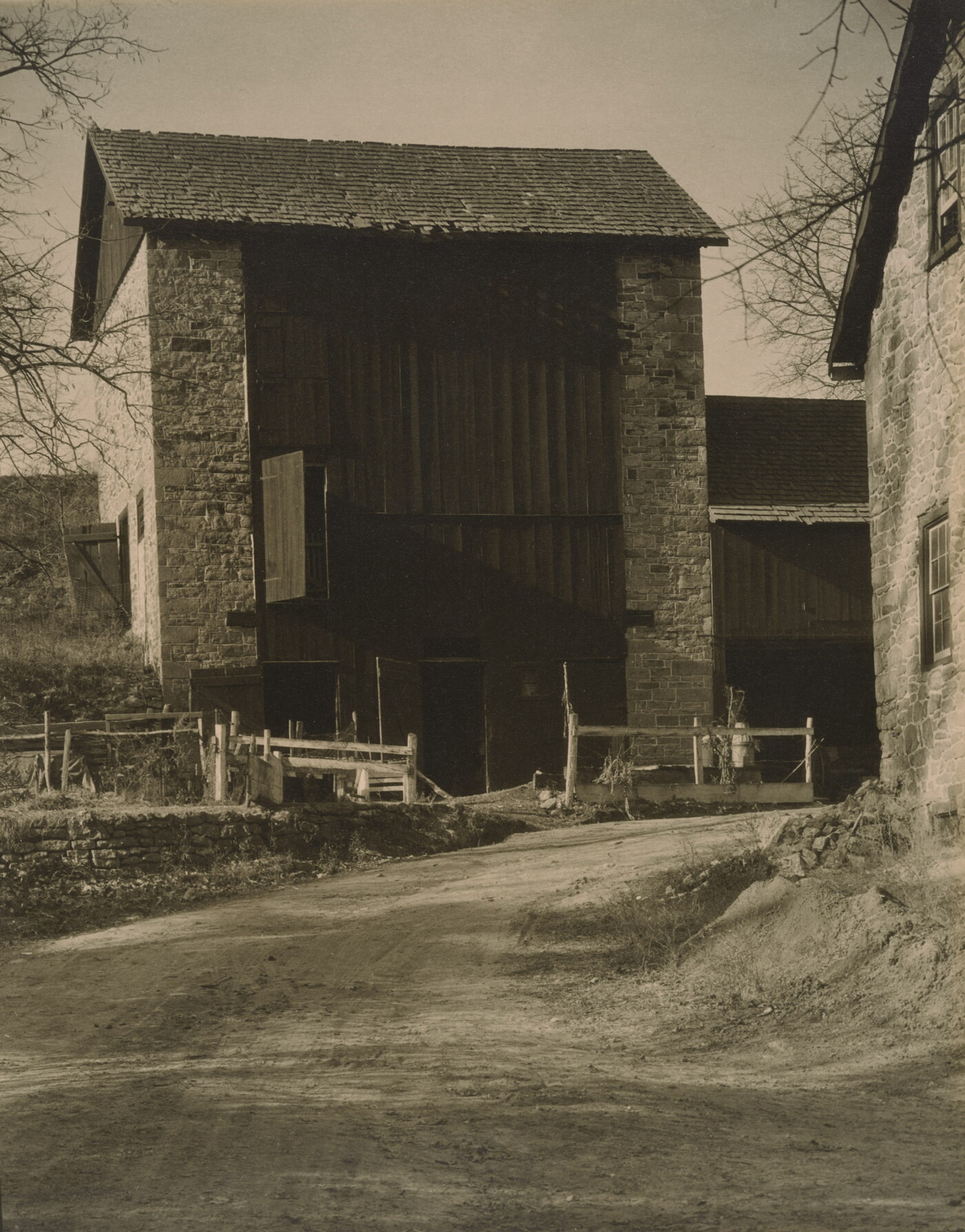 A black-and-white photograph of a dirt road leading to a multi-story stone-sided structure with a wood facade.