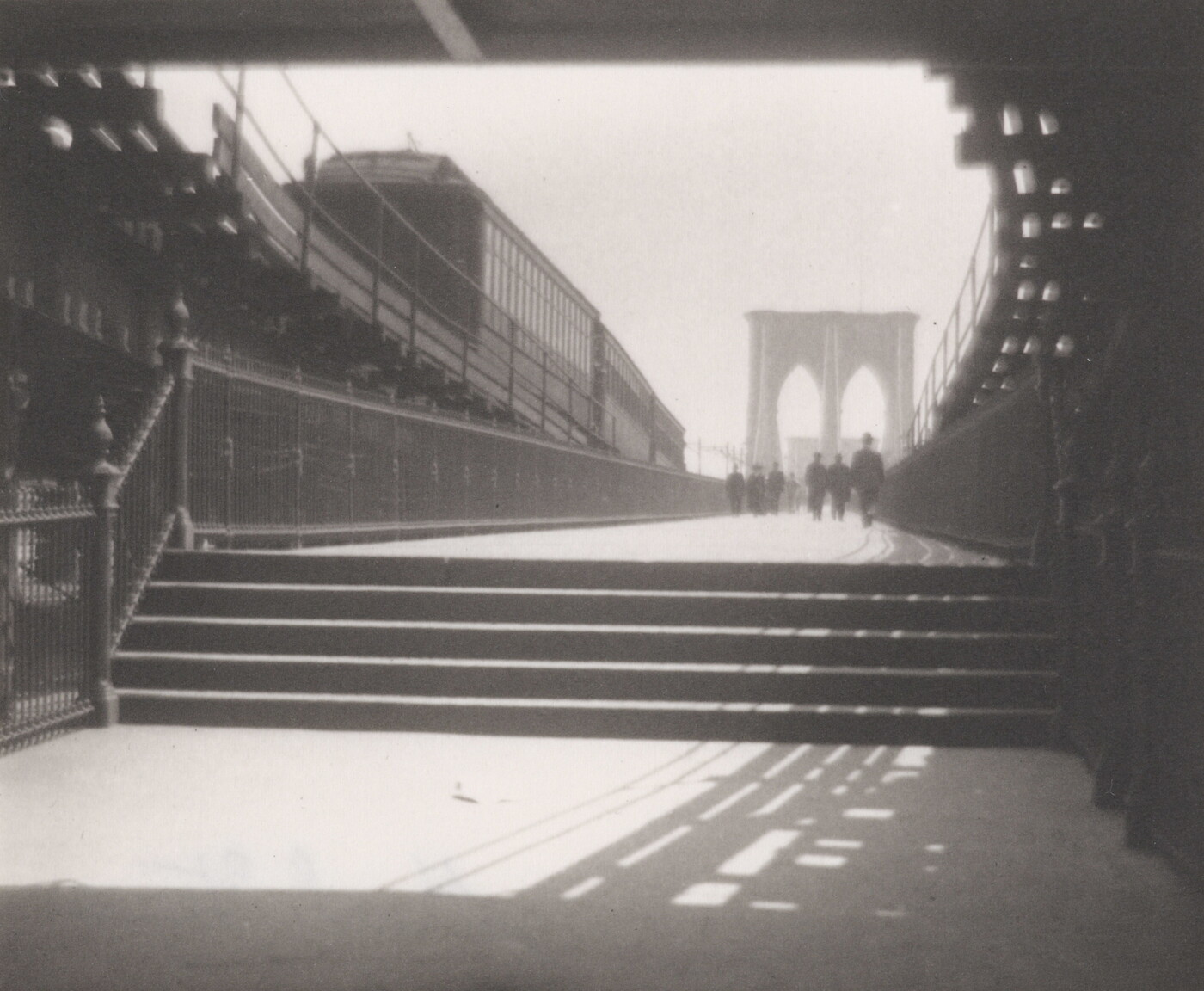 A black-and-white photograph of a set of stairs that leads to the tower of a suspension bridge and people walking toward the tower.