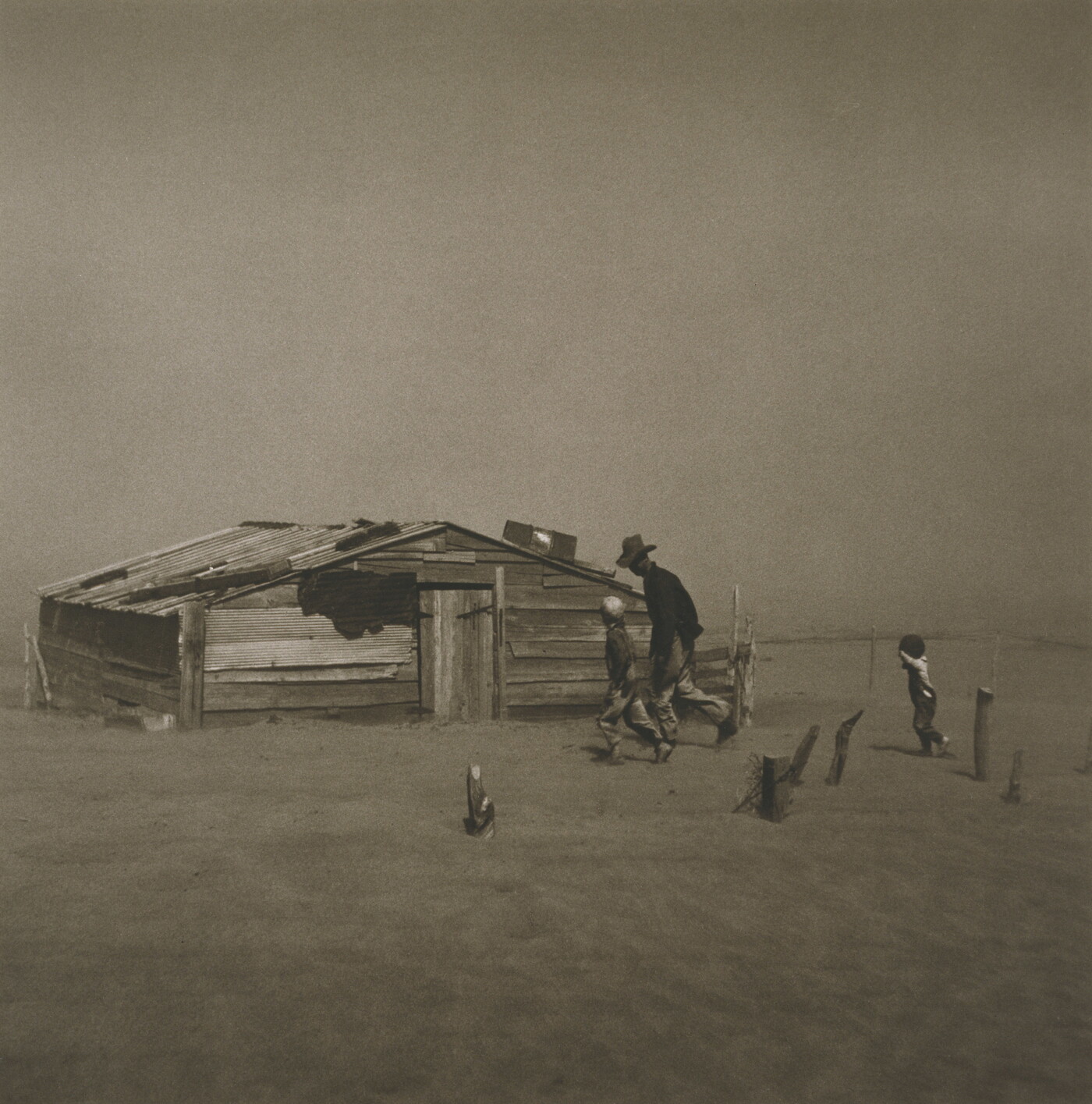 A black-and-white photograph of a man and two children walking through a dust storm by a dilapidated structure.