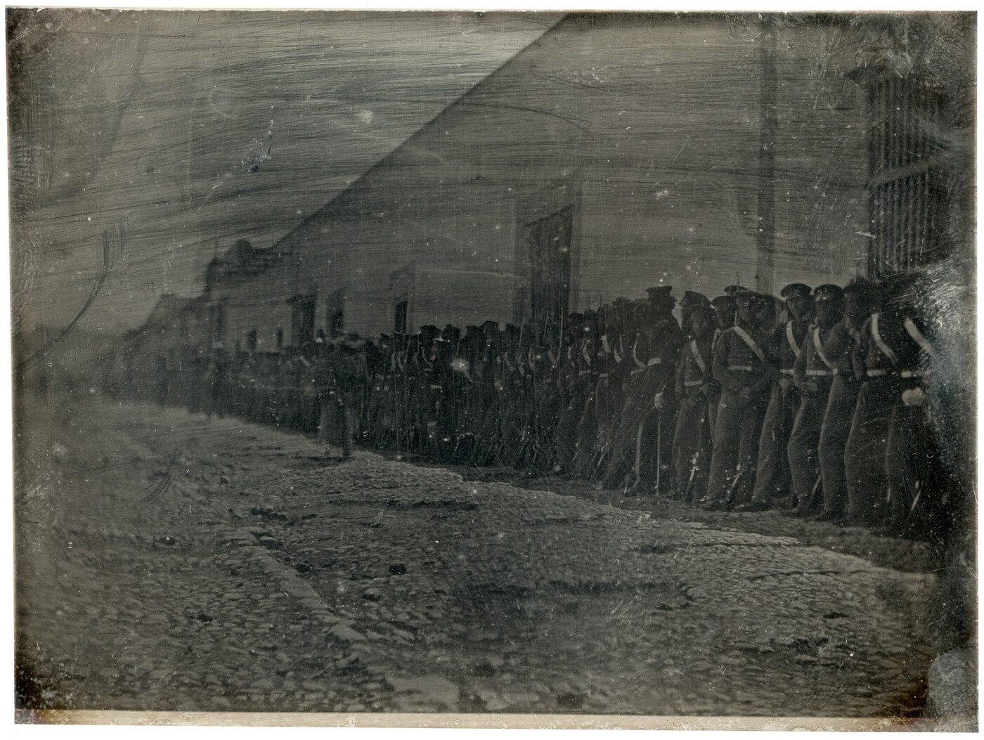 A dark, black-and-white photograph of a group of figures in military uniforms lined up against a building on a cobblestone street.