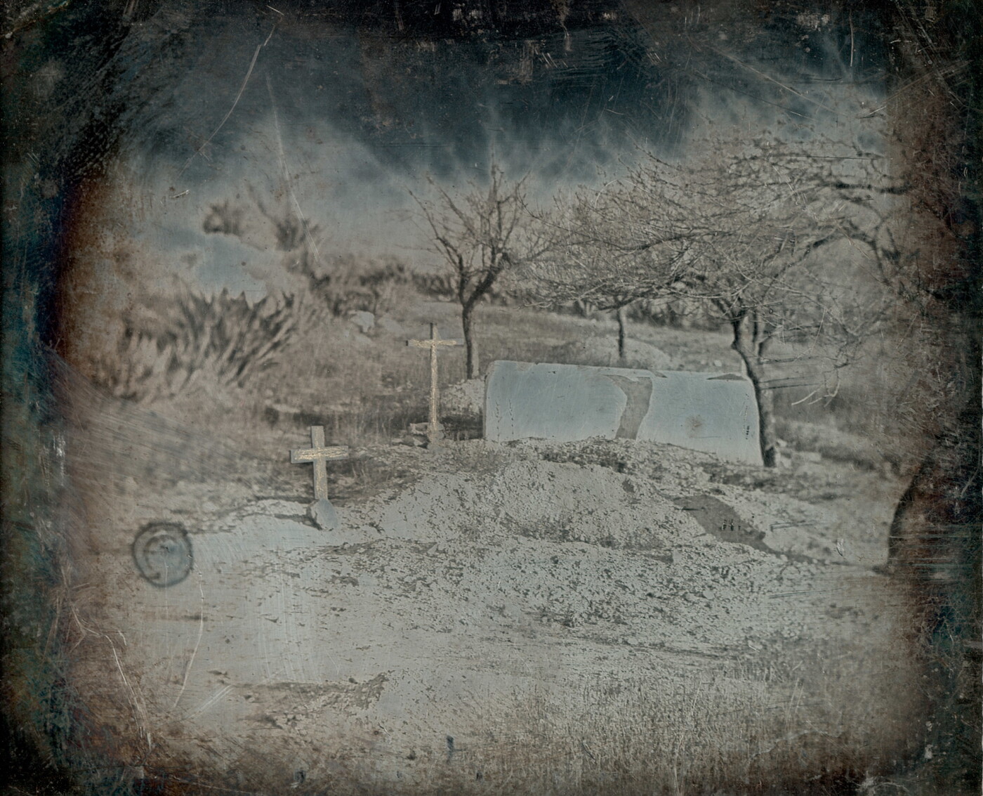 A faded black-and-white photograph of two wooden crosses marking burial sites, surrounded by leafless trees on a dirt hill.