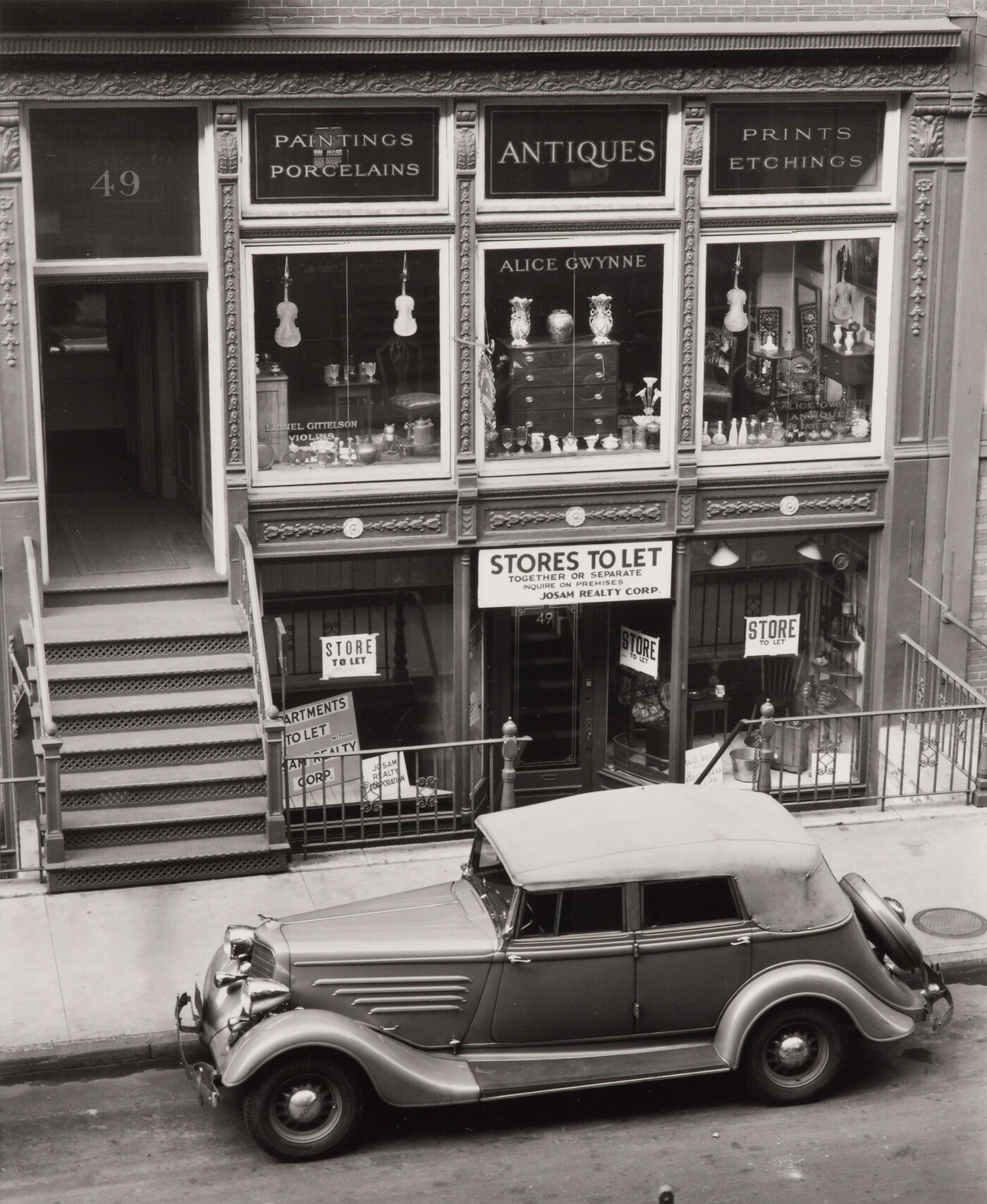 A black-and-white photograph of a vintage car parked on the street outside of a storefront advertising antiques.