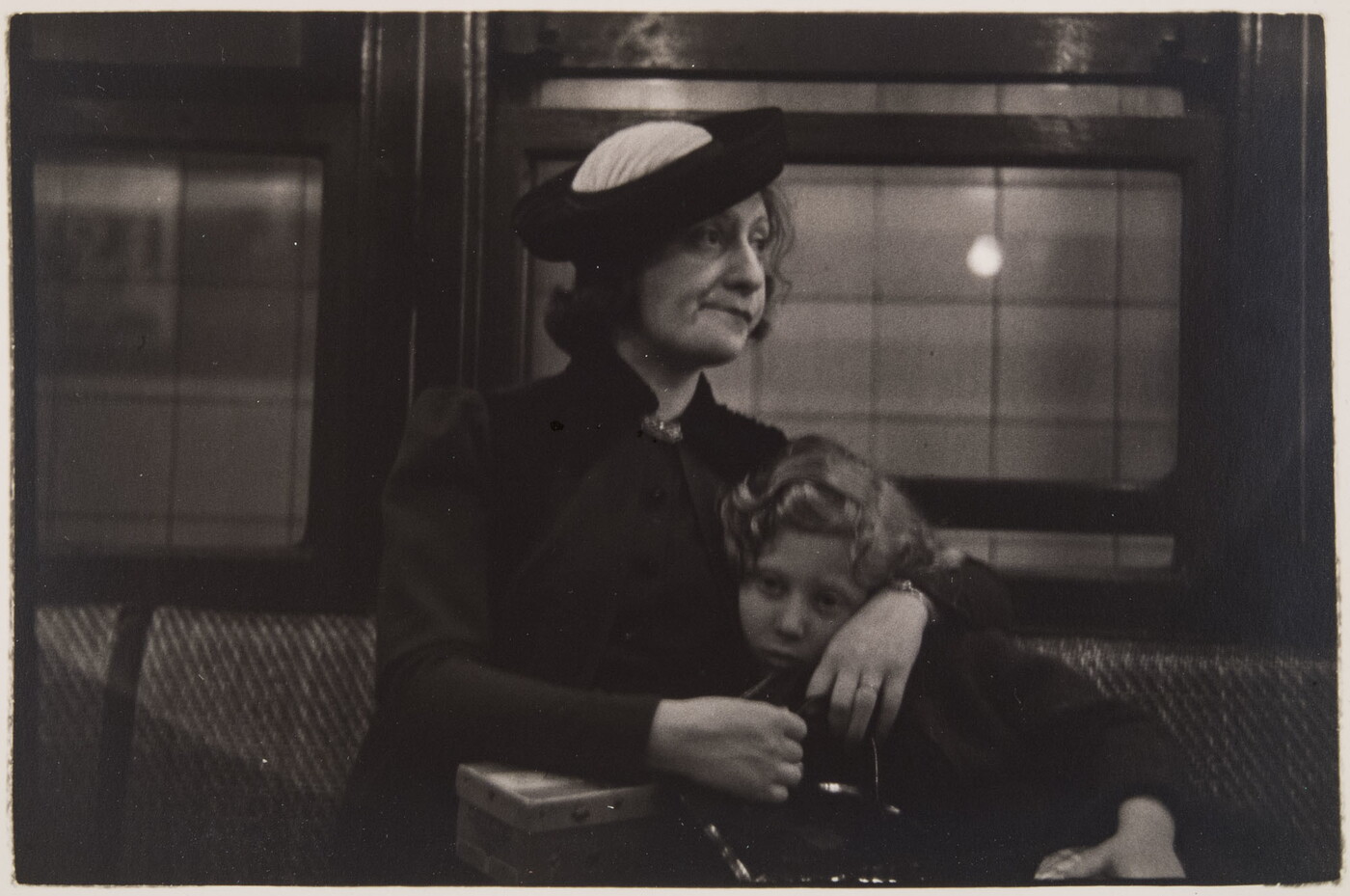 A black-and-white photograph of a White woman with her arms around a child; both seated on an upholstered seat with windows behind them.