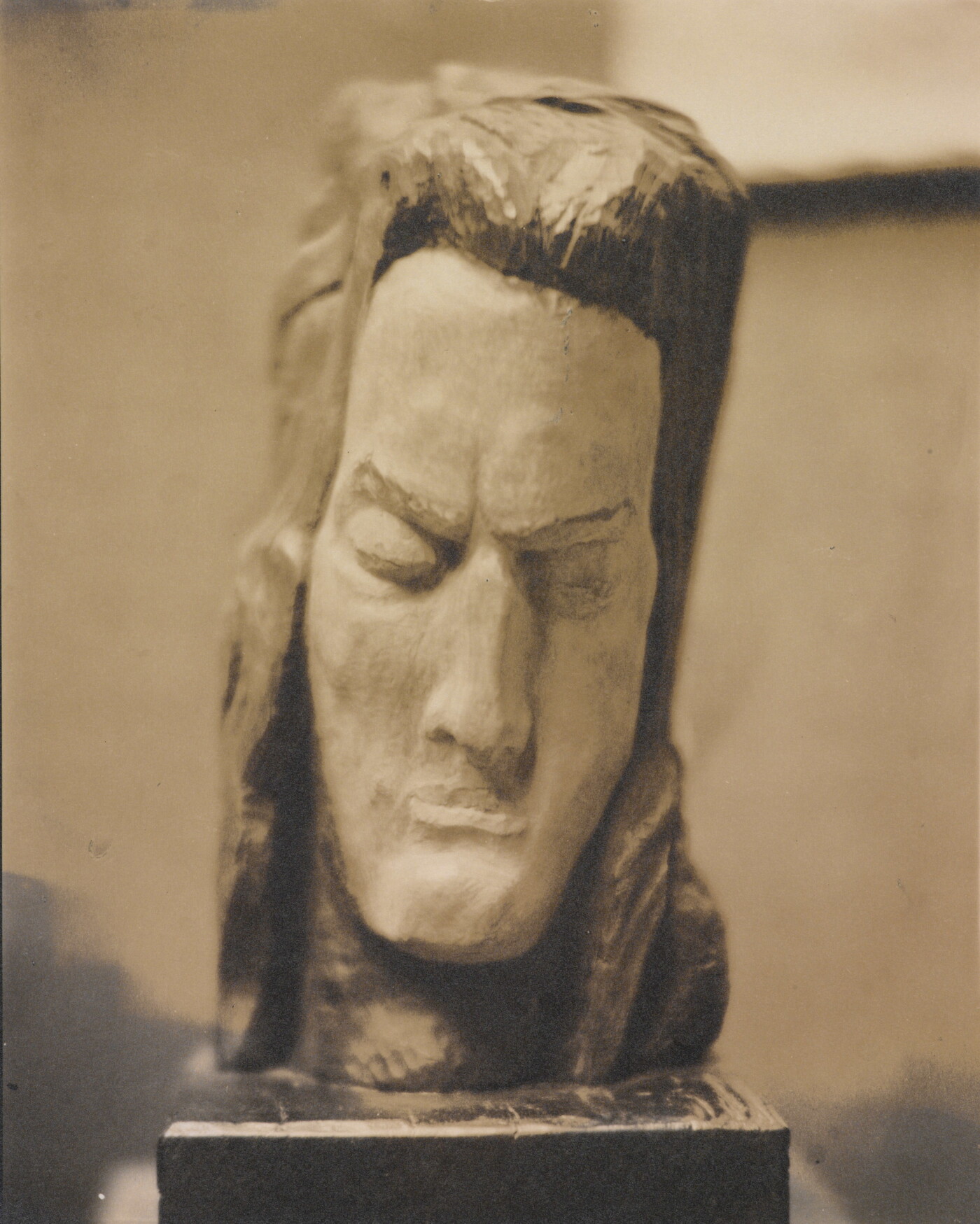 A sepia-toned photograph of a rough-hewn sculpture of a man&#039;s head resting on a pedestal.