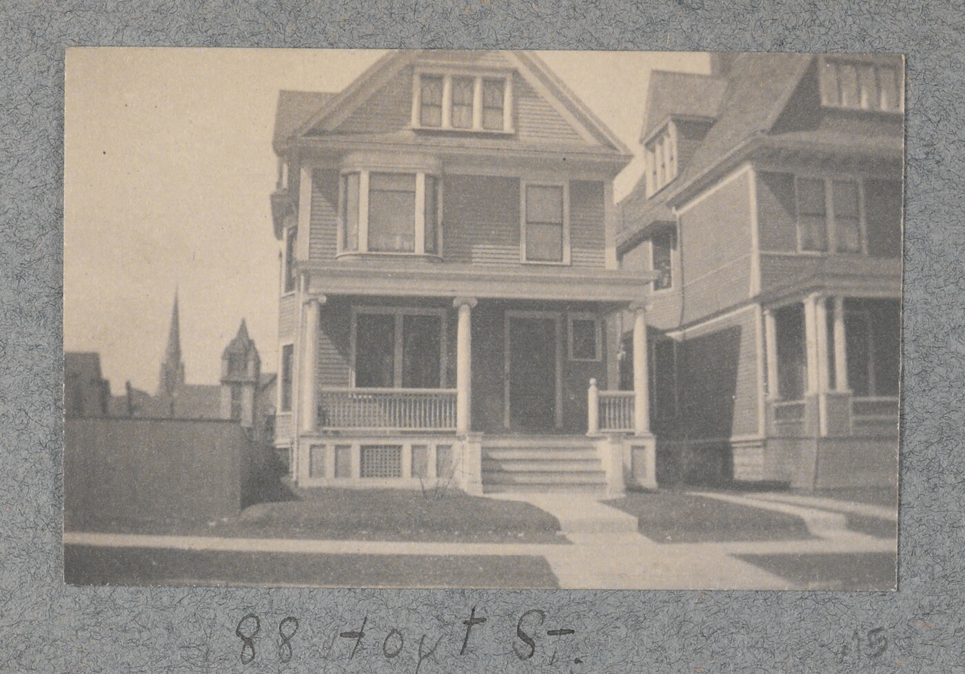 A black-and-white photograph of a three-story wood house, with a front porch and steps leading to a sidewalk in front.