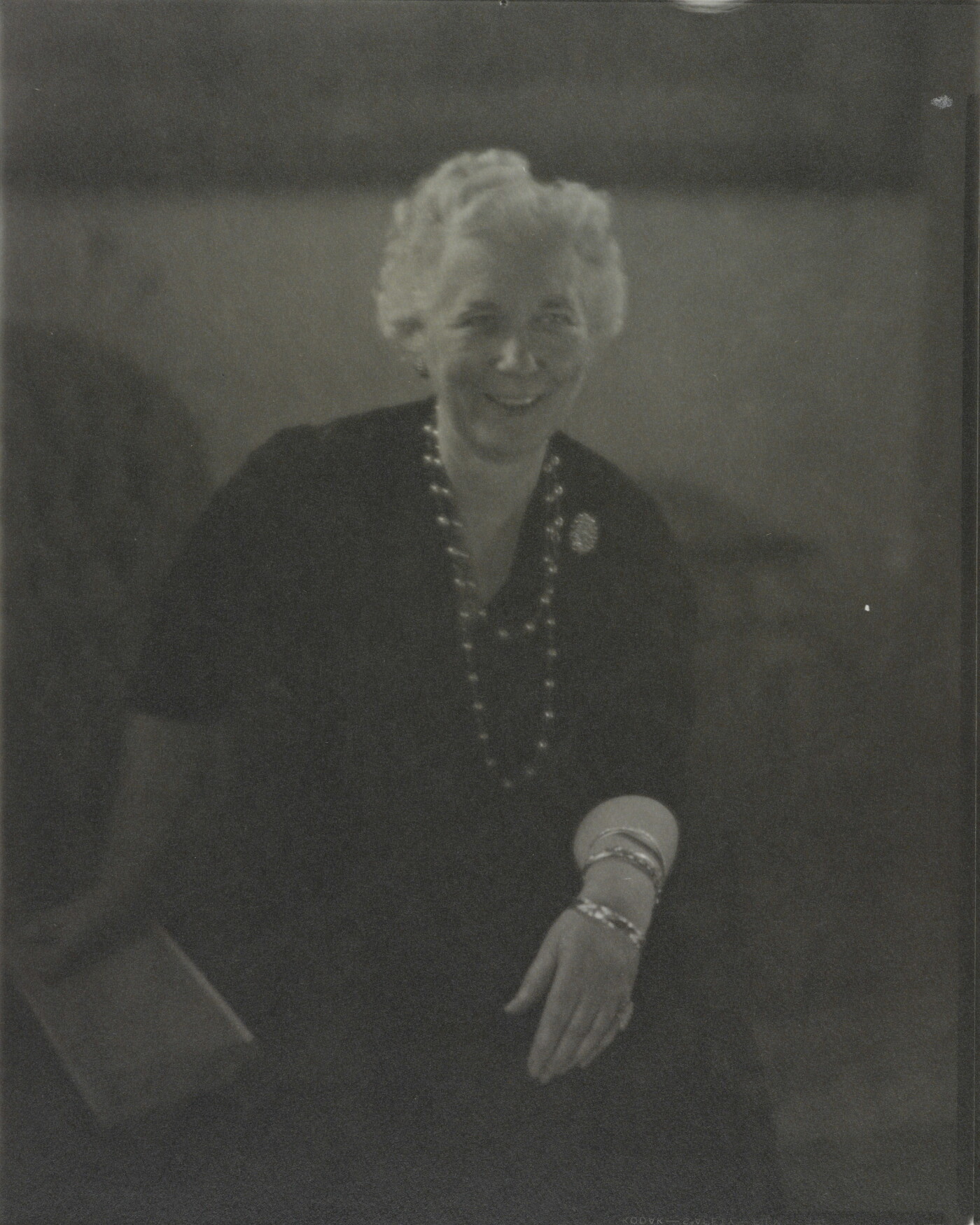 A black-and-white photograph of a smiling White woman with short white hair, seated; one hand holds a book, the other rests on her knee.