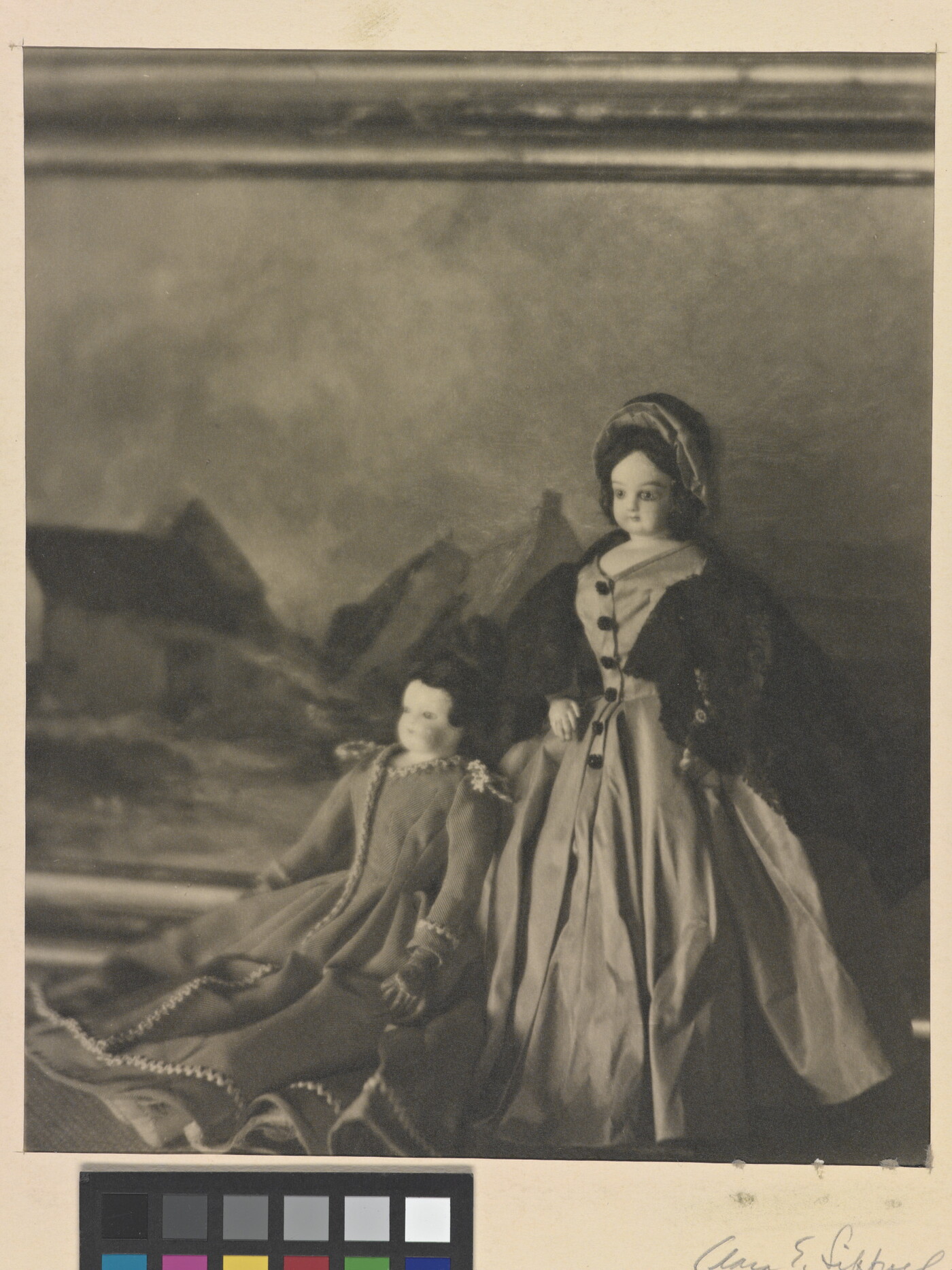 A black-and-white photograph of two light-colored dolls wearing formal 19th century European dresses posed in front of a landscape painting.