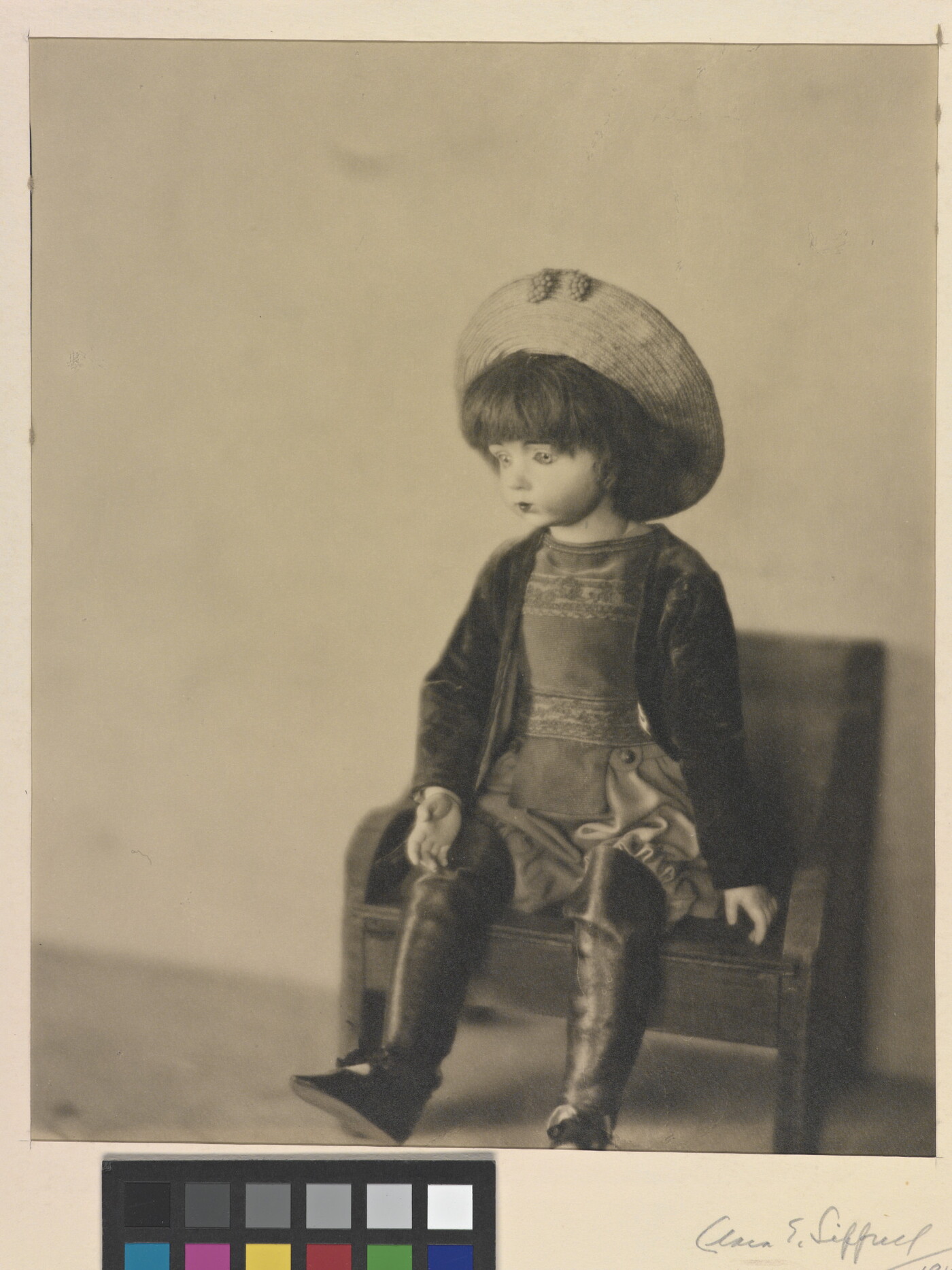 A sepia-toned photograph of a light-colored doll wearing a hat and boots sitting on a small bench.