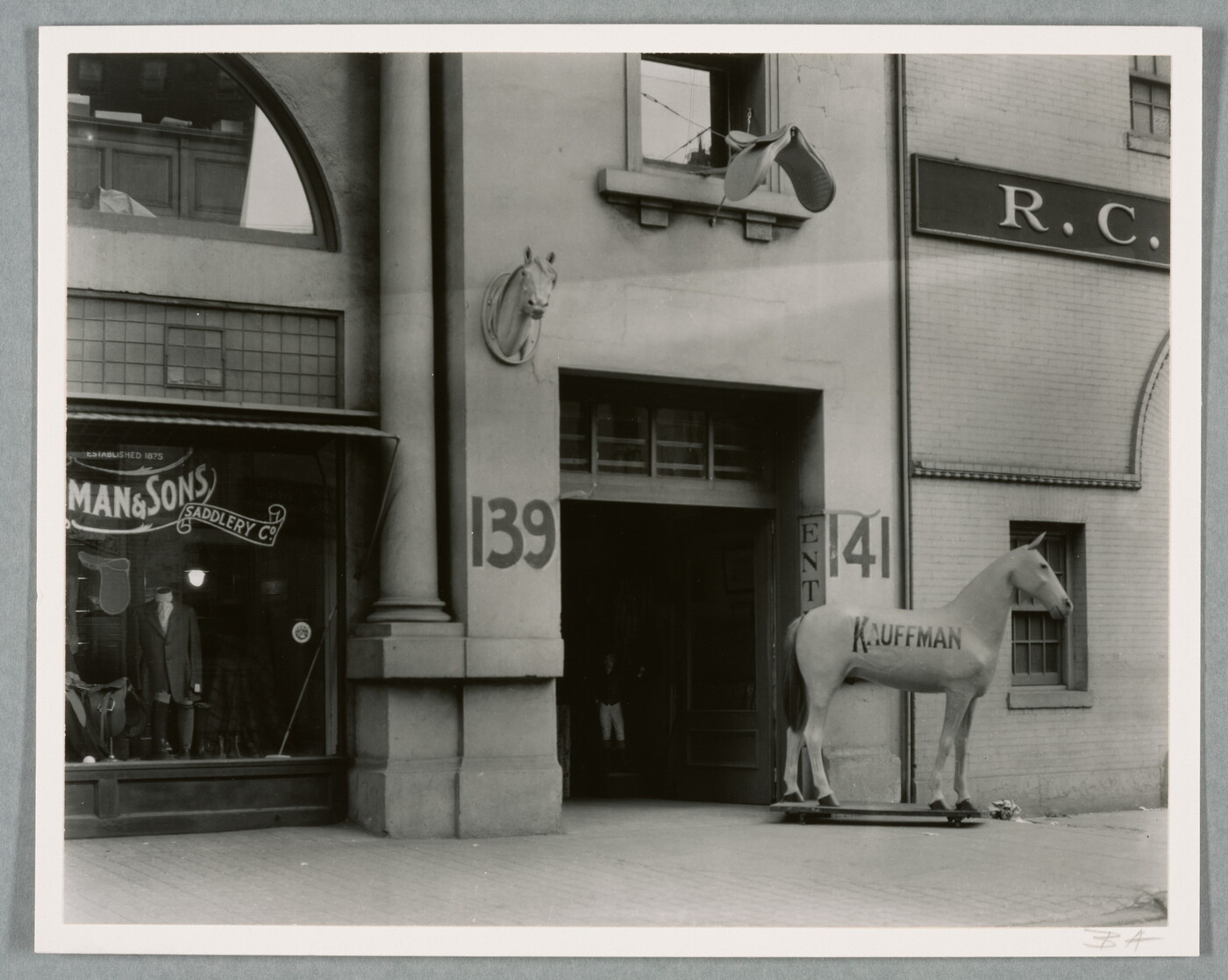 A black-and-white photograph of a storefront with a large statue of a horse, the word "Kauffman" painted on its side, next to the entrance.