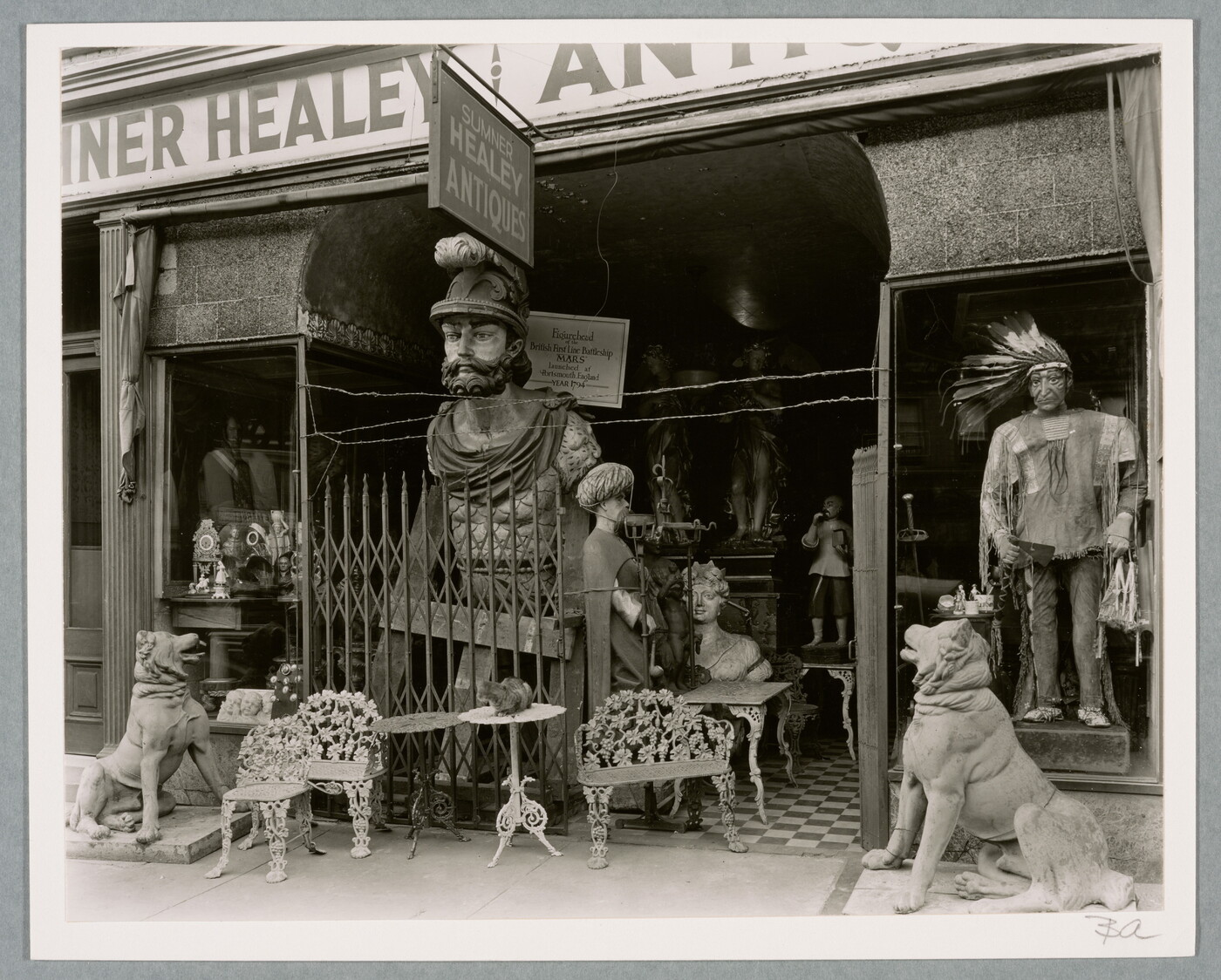 A black-and-white photograph of an overcrowded storefront with a variety of furniture and large statues at the entrance.