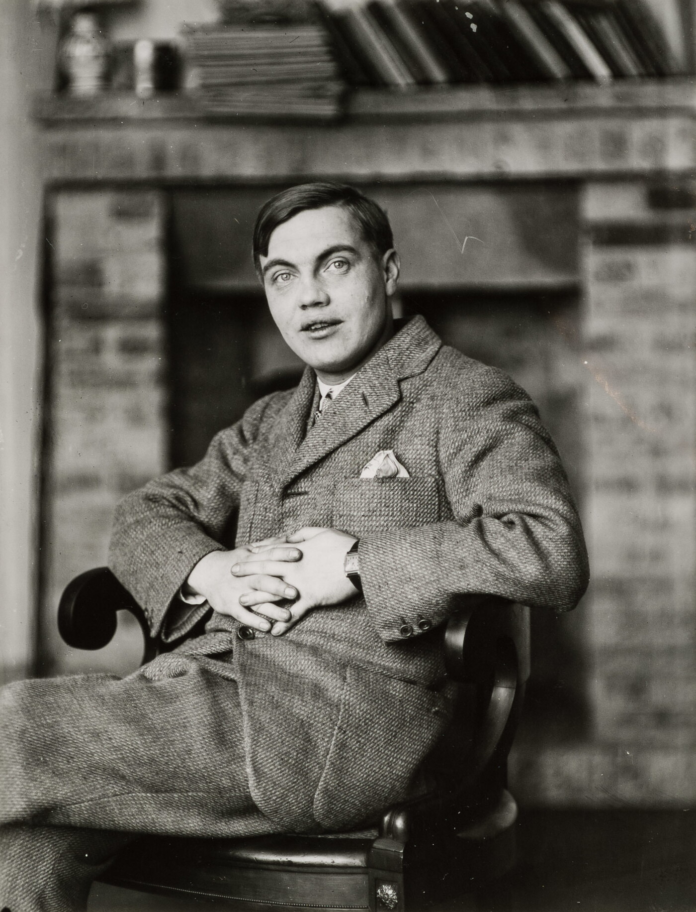 A black-and-white portrait photograph of a White man in a suit sitting in a wood chair with his hands clasped in front of him.