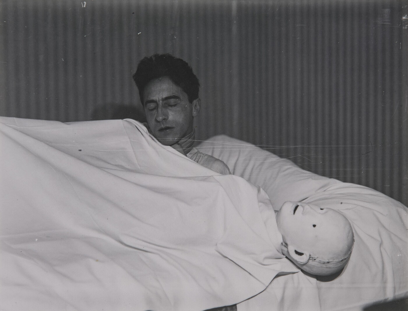 A black-and-white photograph of a White man lying under a white sheet next to a mannequin head.