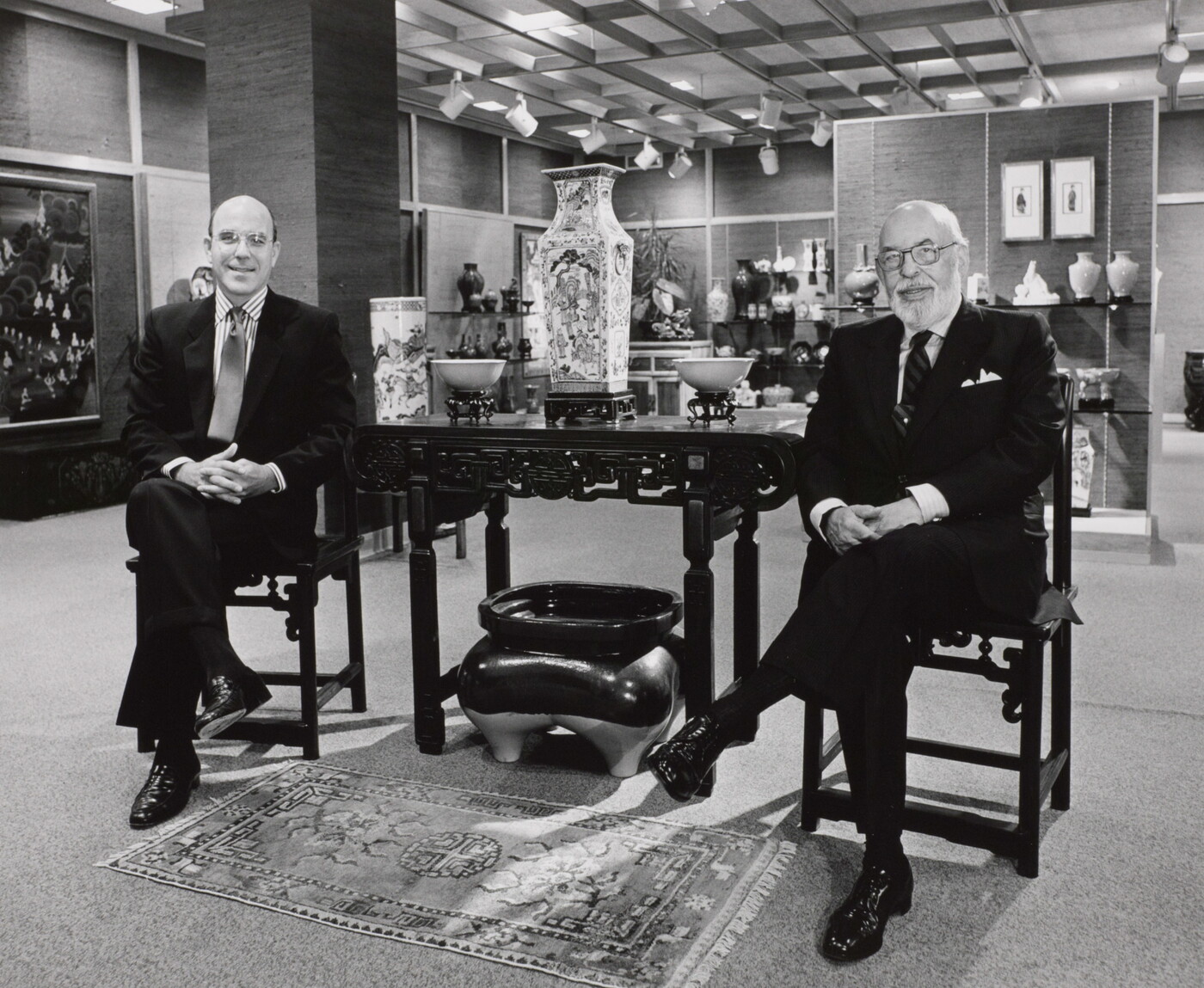 A black-and-white photograph of two smiling White men in suits sitting on either side of a table displaying Asian decor.