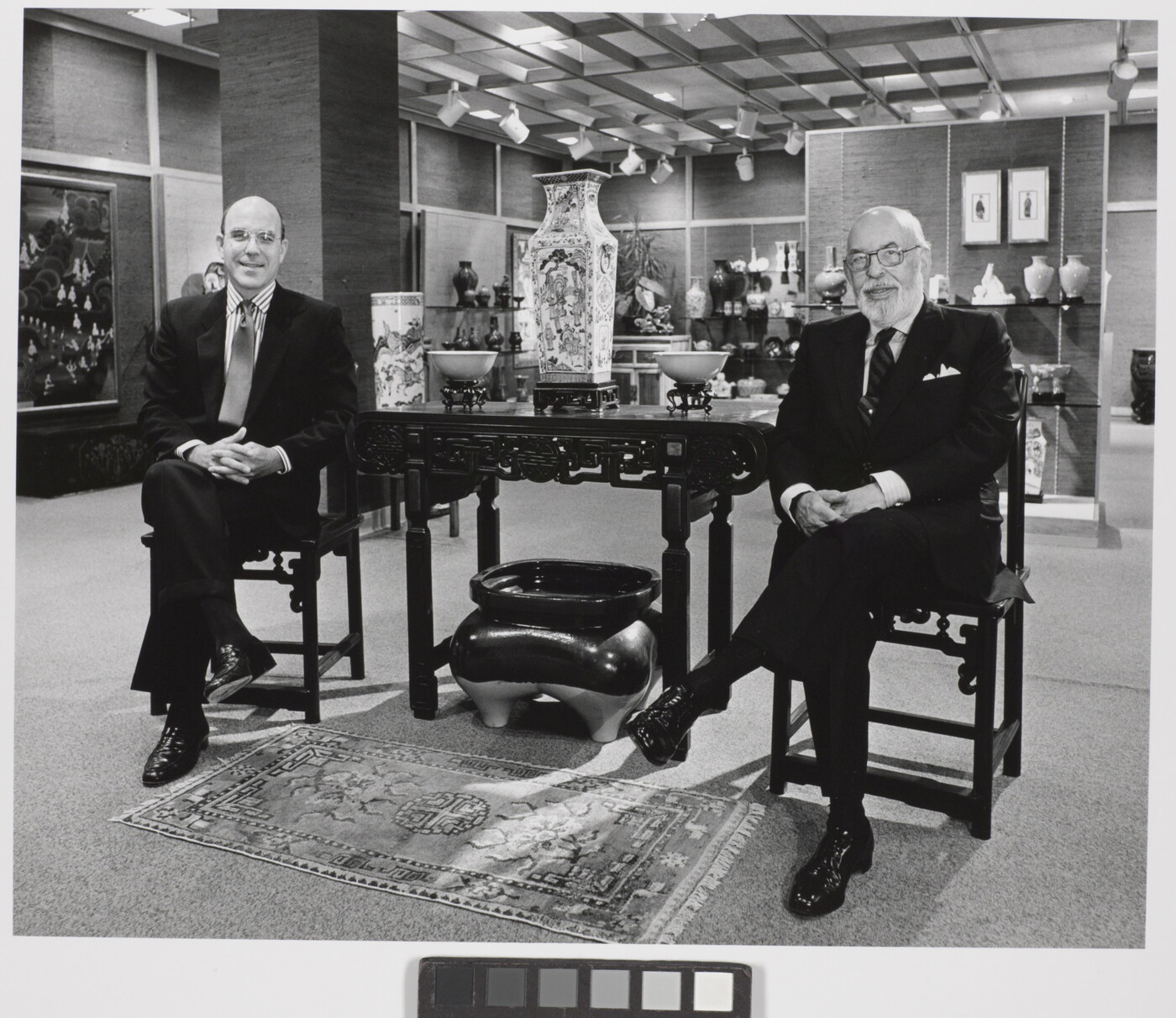 A black-and-white photograph of two smiling White men in suits sitting on either side of a table displaying Asian decor.
