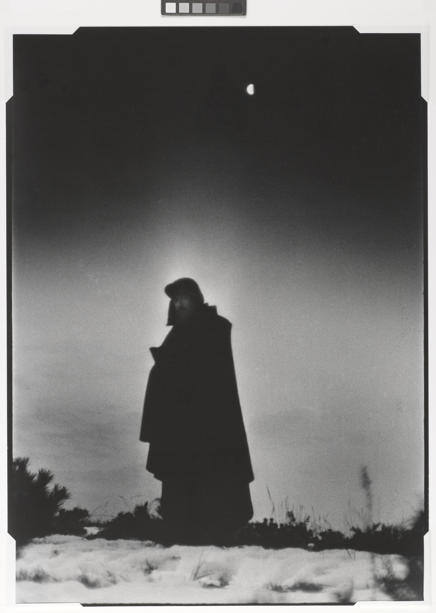 A black-and-white photograph of a heavily cloaked person standing in a frozen landscape with a half-moon behind them.