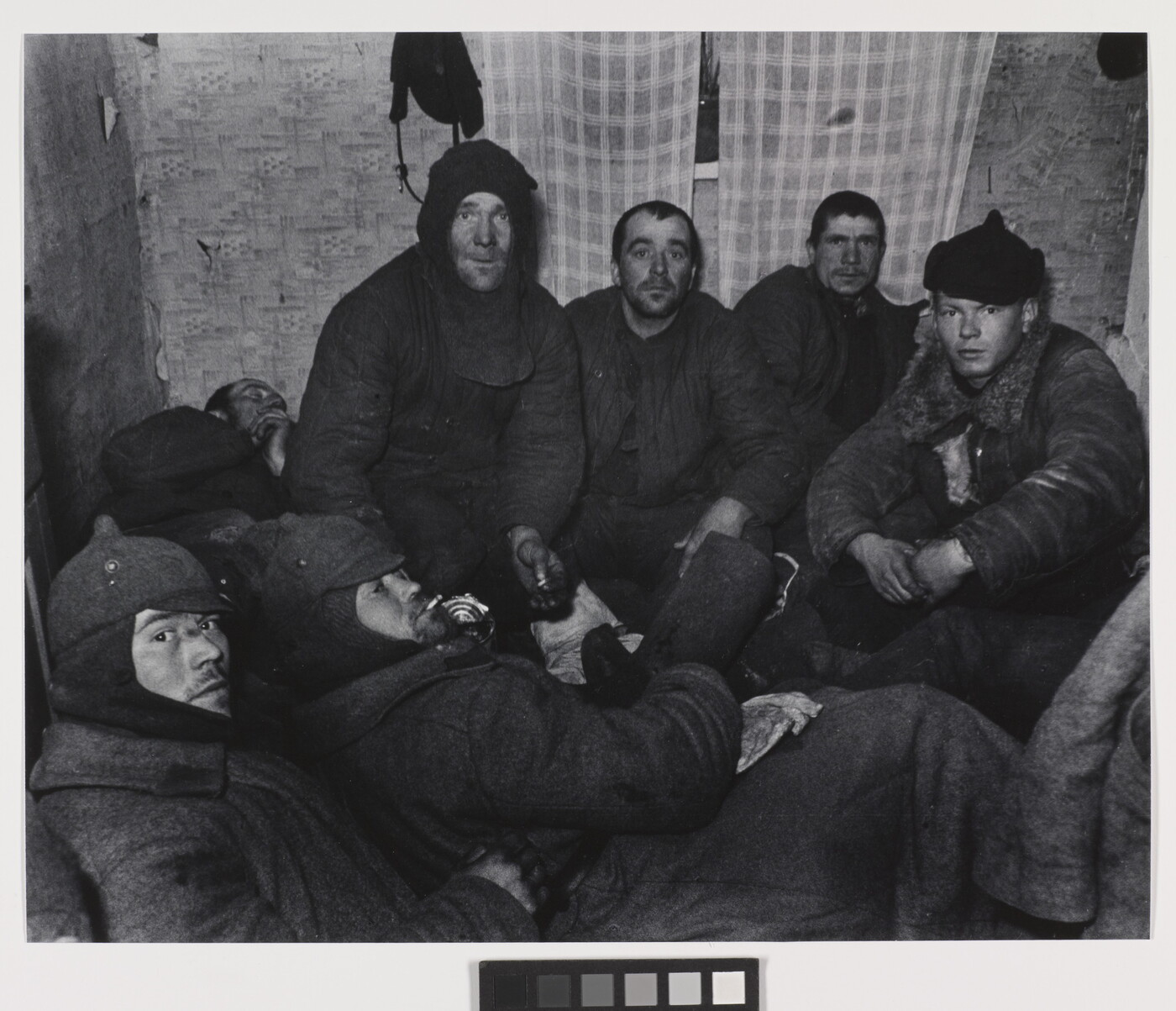 A black-and-white photograph of a group of White men wearing dark, heavy clothing huddled in a small room with plaid curtains over a window.
