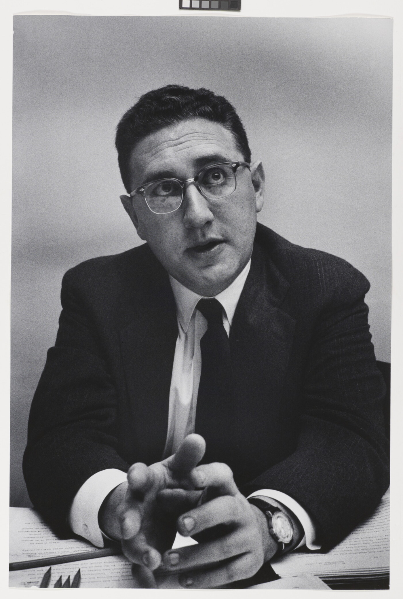 A black-and-white photograph of a White man with short, dark hair in a suit, tie, and glasses leaning forward with his elbows on a desk, gesturing with his hands.