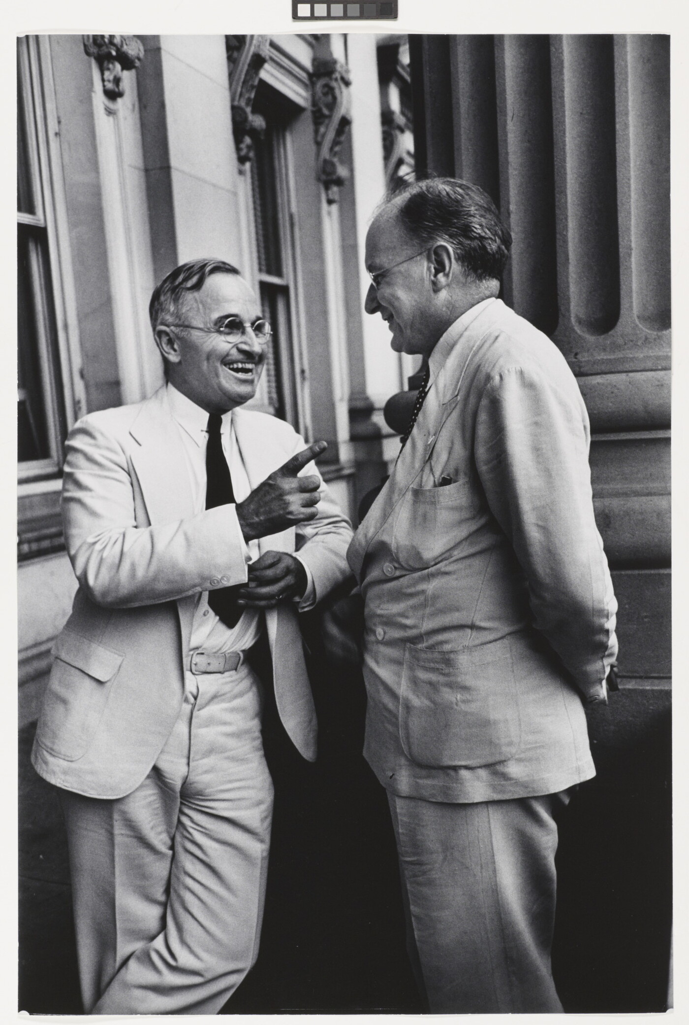 A black-and-white photograph of two White men, both in light-colored suits and glasses, talking to one another and smiling.