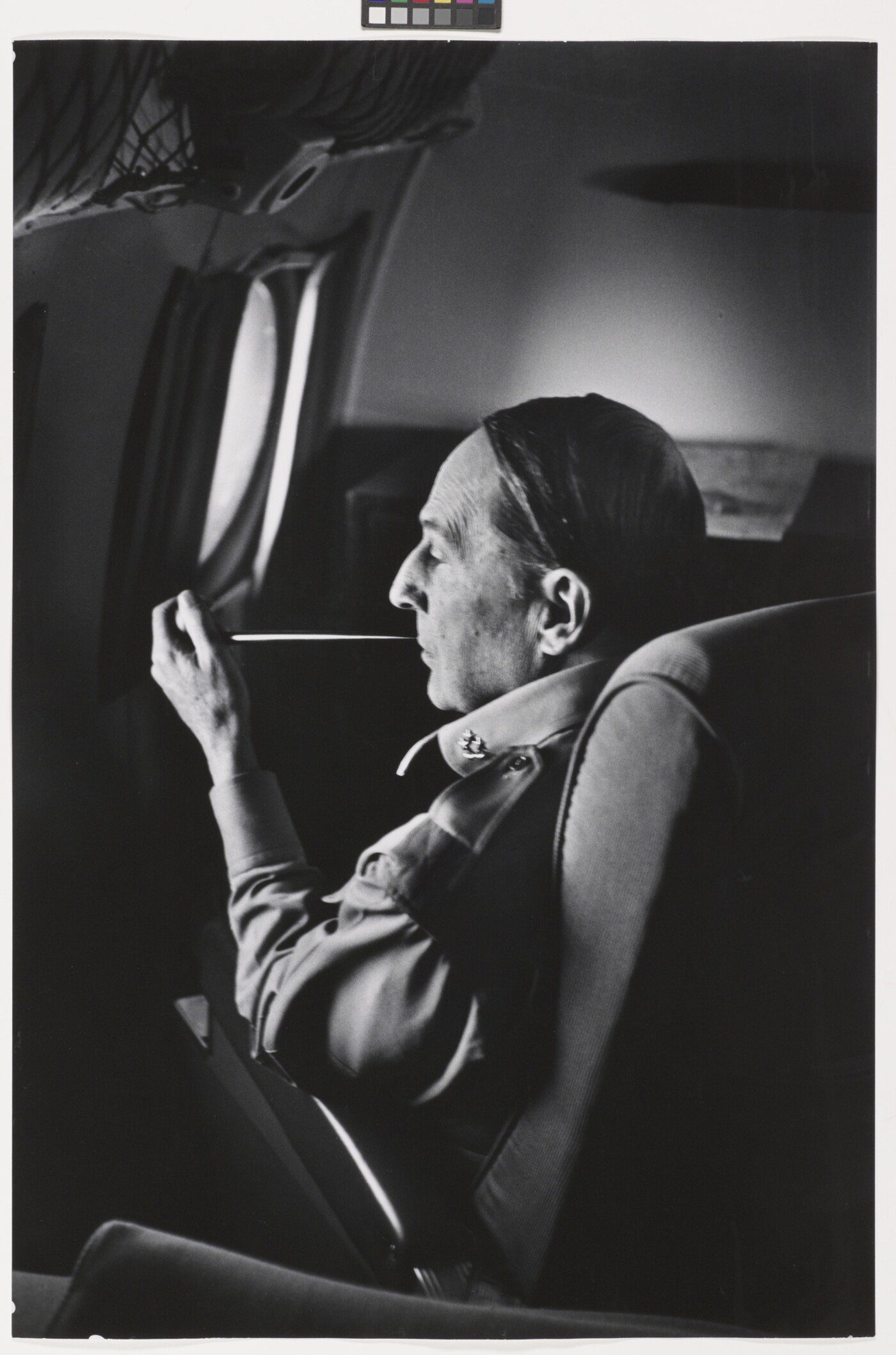 A black-and-white photograph of a White man seated on an airplane smoking a long thin pipe.