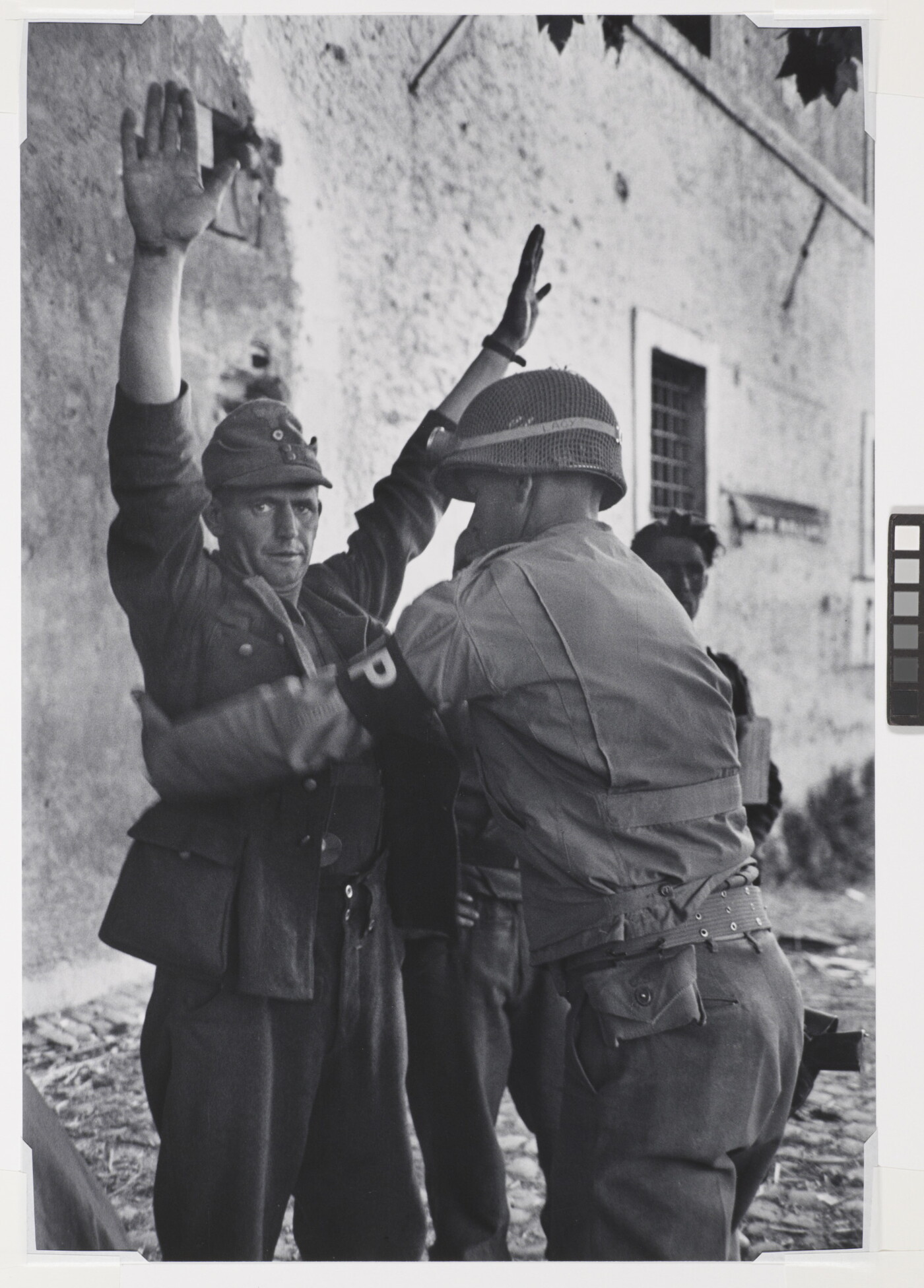 A black-and-white photograph of a White man in military uniform being frisked by a second man in military uniform.