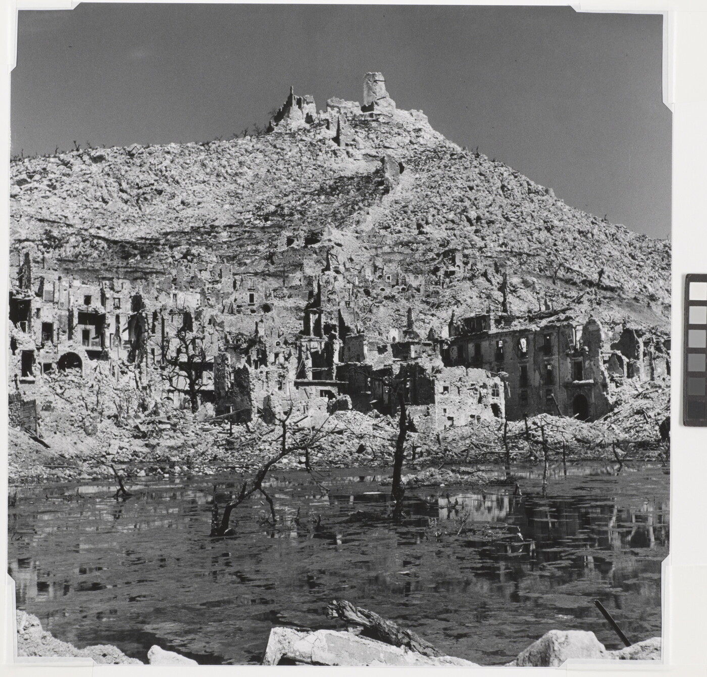 A black-and-white photograph of the ruins of a town at the edge of a body of water and a ruined building on top of a mountain in the background.