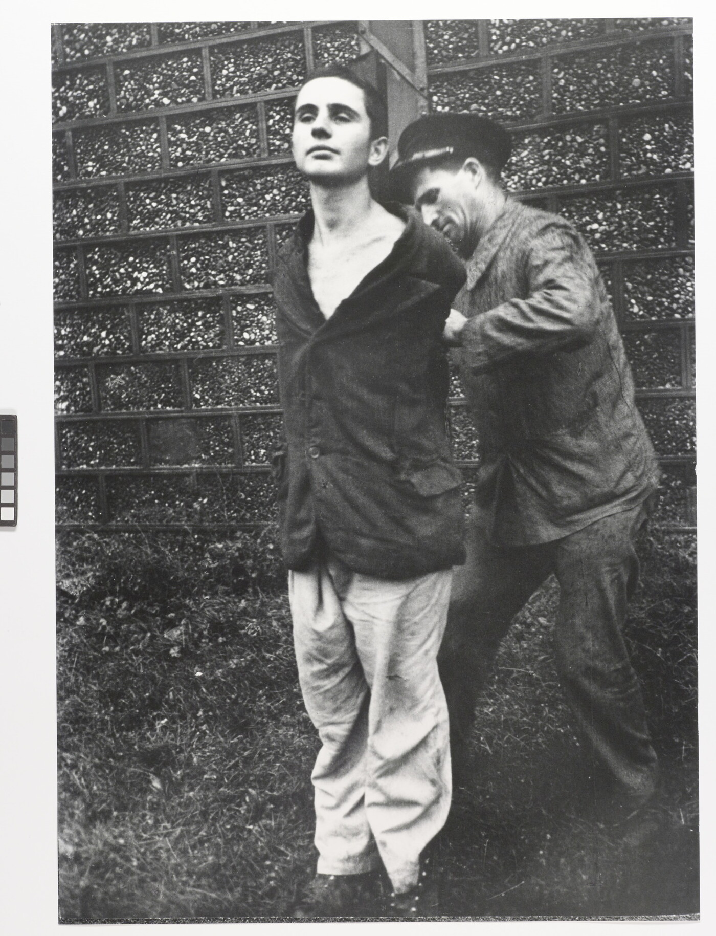 A black-and-white photograph of a young, White man standing with his hands behind his back as another White man ties them to a wood pole.