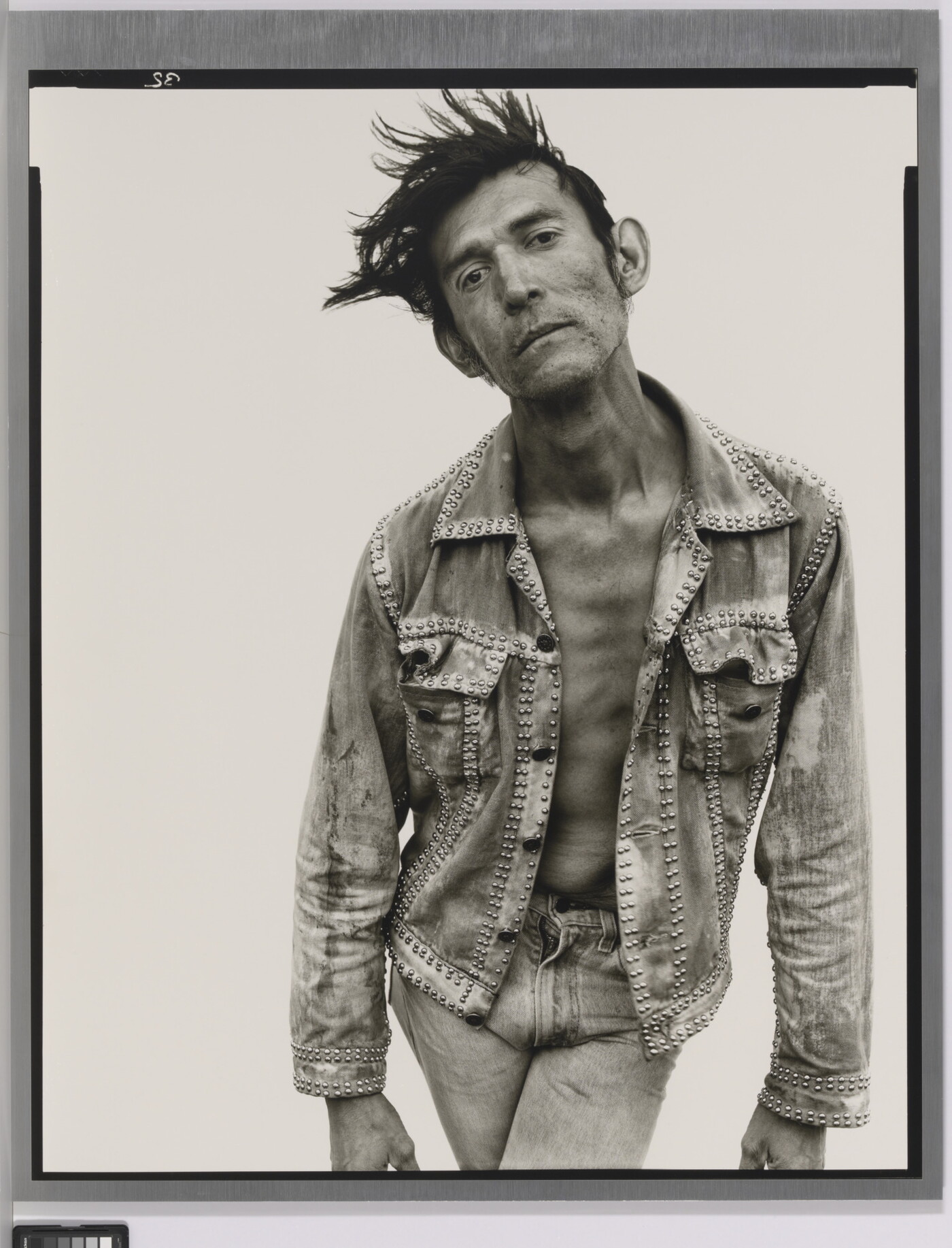 A black-and-white portrait photograph of a White man with dark hair that sticks up, wearing jeans and a jean jacket, and crossing his legs while bending forward.