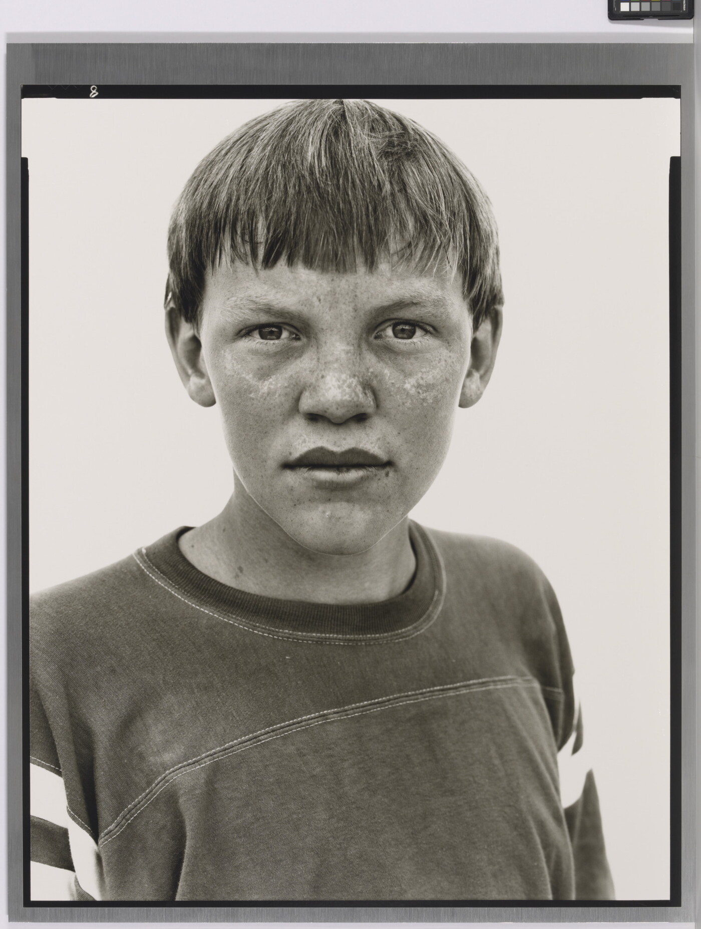 A black-and-white portrait photograph of a White teenage boy with a bowl haircut and freckles across his nose and cheeks, wearing an athletic t-shirt.