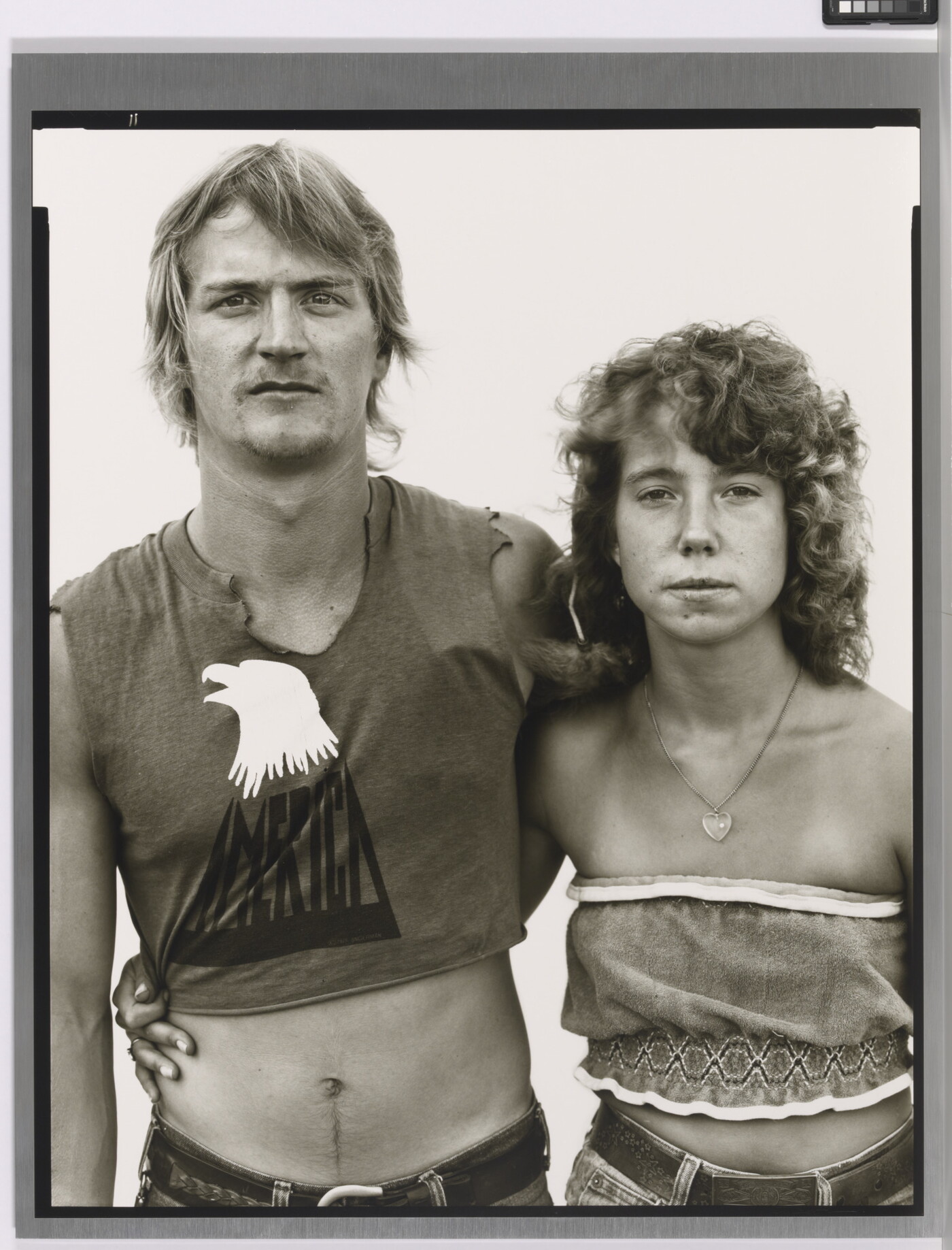 A black-and-white portrait photograph of a young White man and woman, arms around each other, both wearing crop-top shirts.