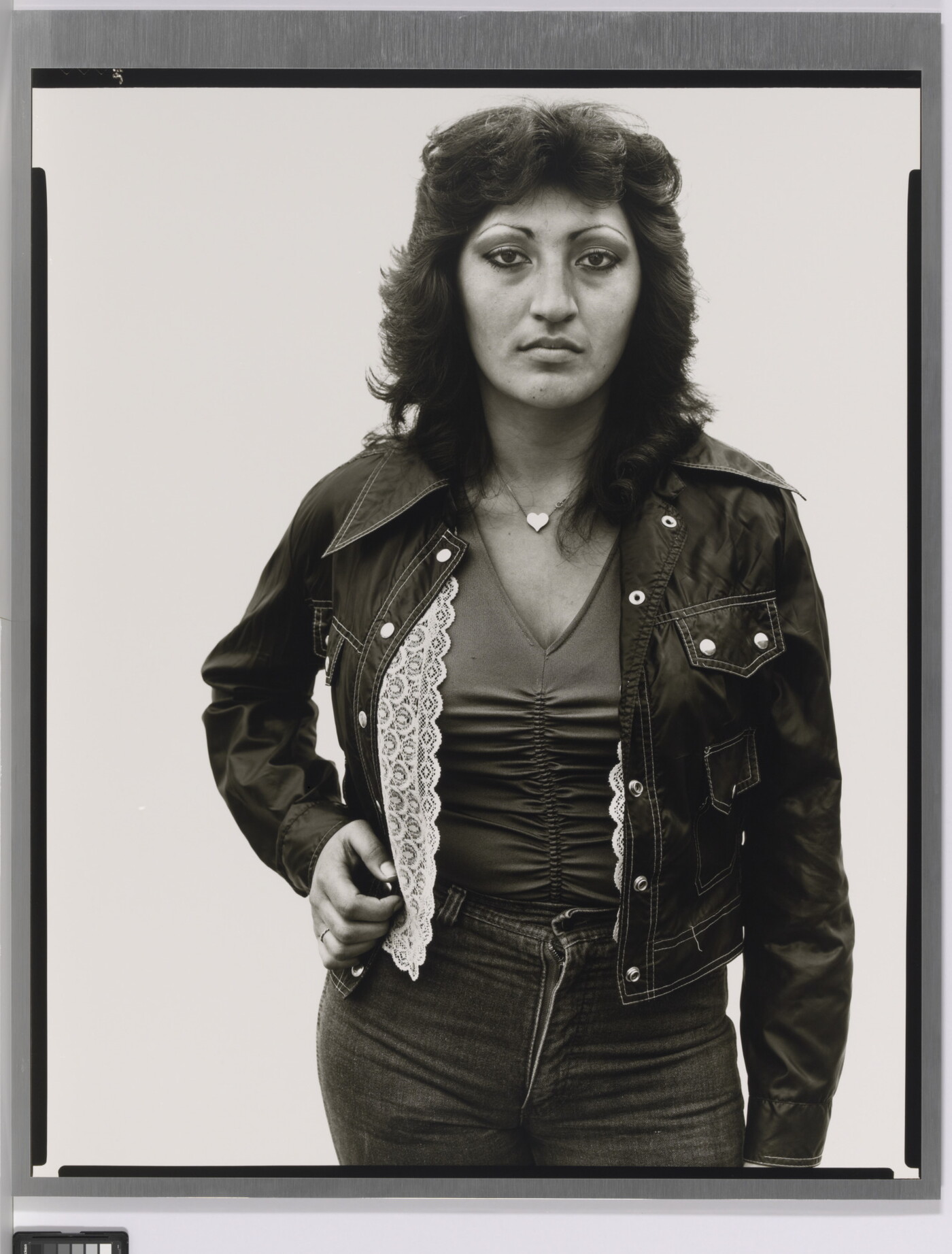A black-and-white portrait photograph of a medium-skinned woman with dark, feathered hair wearing jeans, a jacket, V-neck top, and a heart necklace.