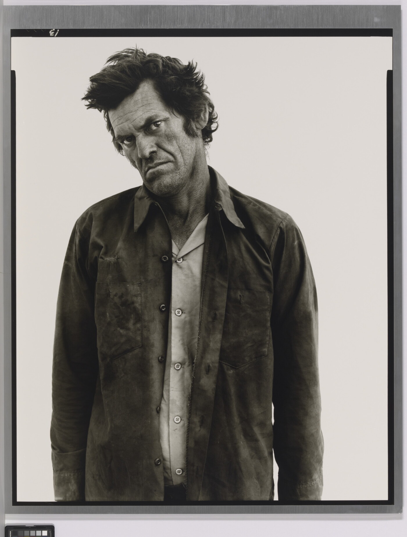 A black-and-white portrait photograph of a White man standing with his head cocked, frowning, and wearing two soiled shirts.