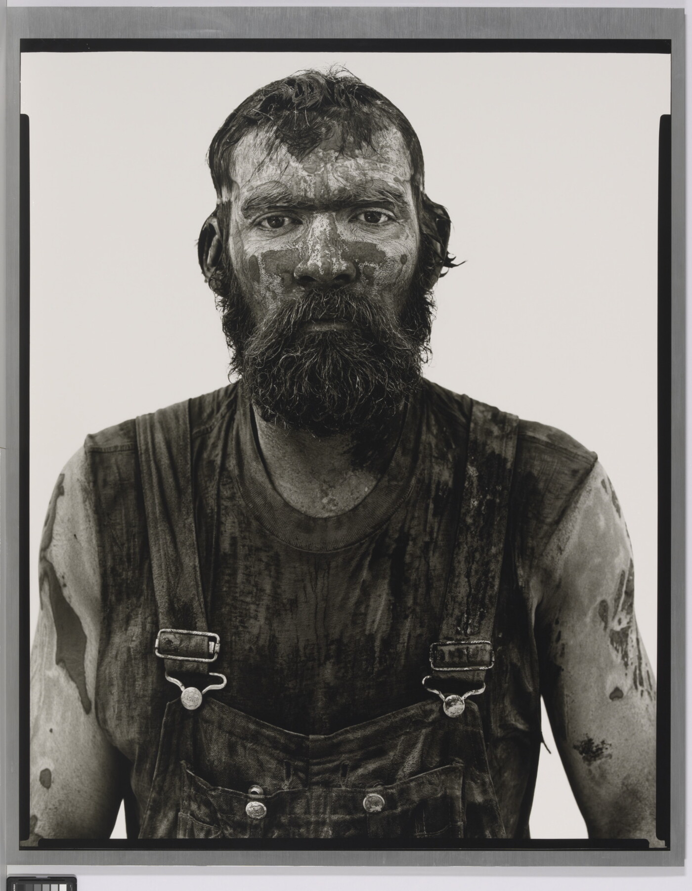 A black-and-white portrait photograph of a bearded man covered in mud and dirt, wearing dirty overalls, and looking straight at the camera.