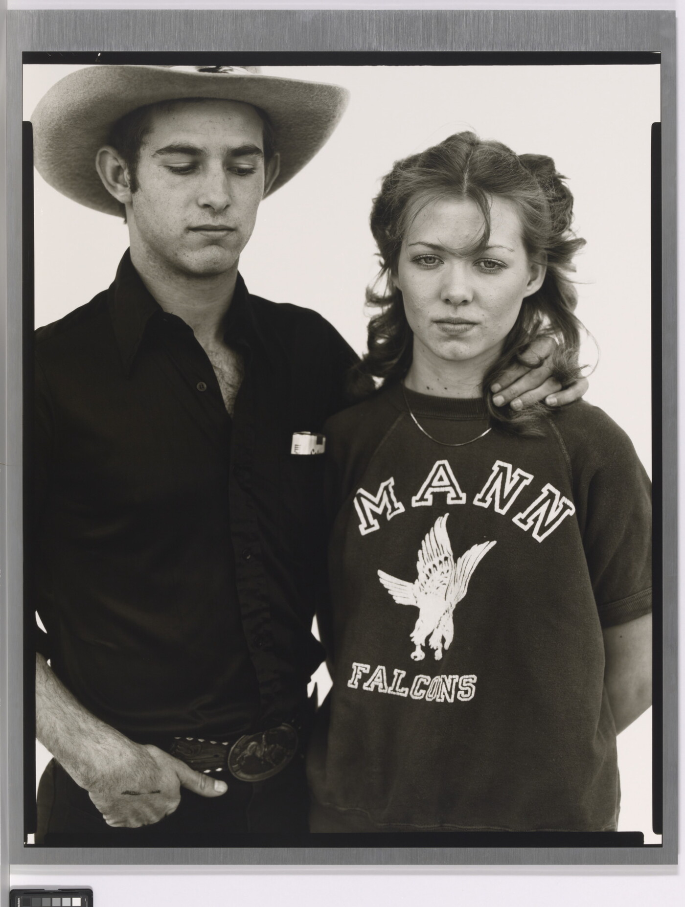 A black-and-white portrait photograph of a young White man wearing a cowboy hat, looking down, his hand affectionately around the neck of a young White woman.