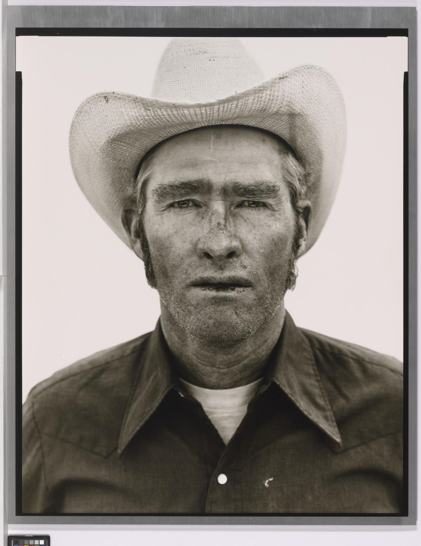 A black-and-white portrait photograph of a White man wearing a cowboy hat and collared shirt, with stubble on his face and chapped lips.